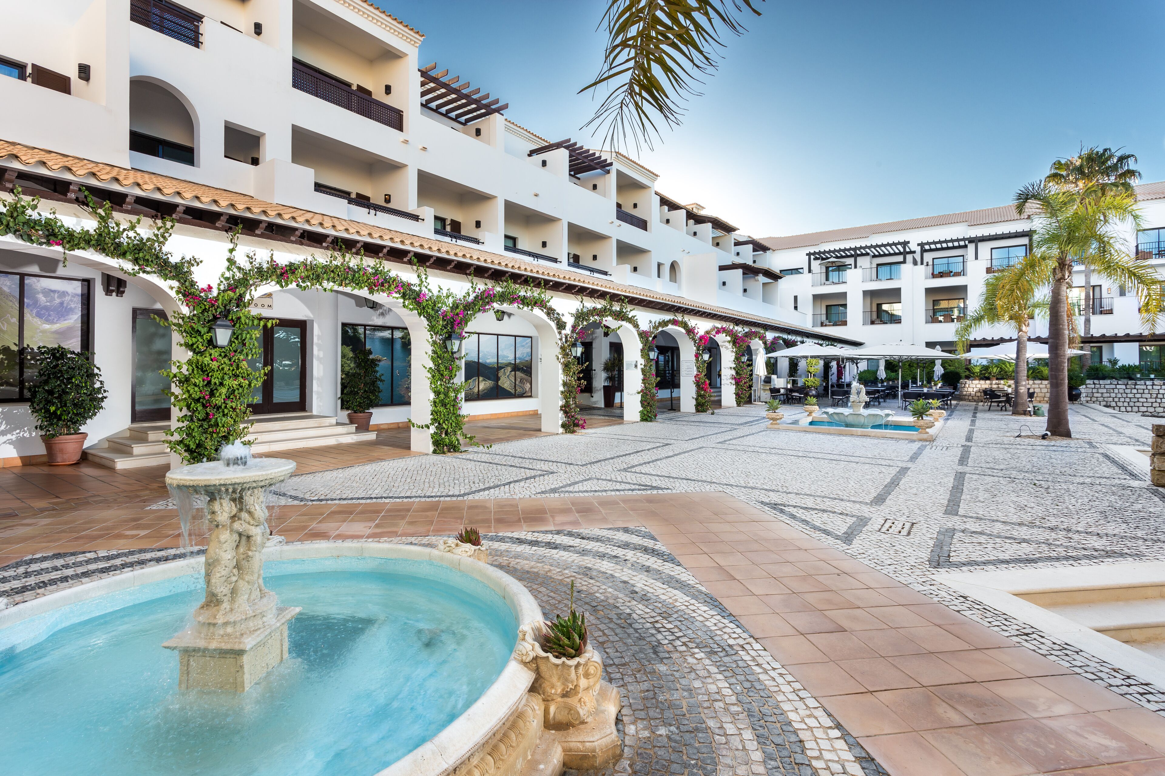 A courtyard with a fountain, tiled flooring, and a building with multiple balconies and arches.