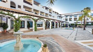 A courtyard with a fountain, tiled flooring, and a building with multiple balconies and arches.