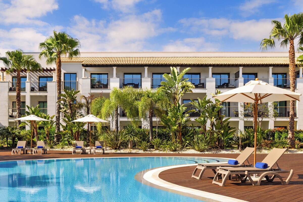 Un espace piscine d’hôtel avec des chaises longues et des parasols, entouré de palmiers et d’un bâtiment doté de balcons.