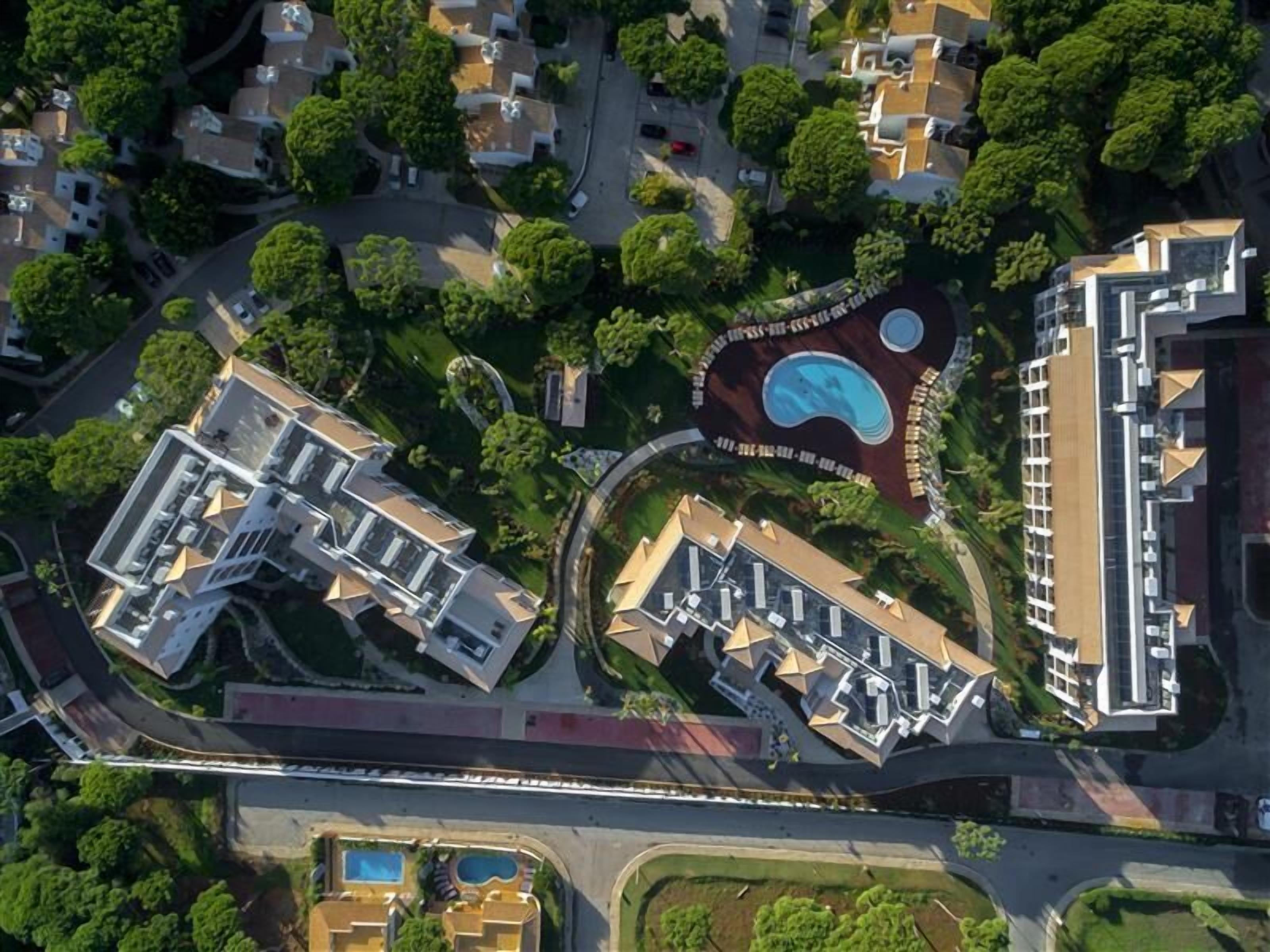 An aerial view of a residential area with modern buildings, a central circular park with a swimming pool, and a connecting road.