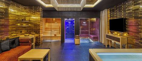 A modern spa room with a seating area, a massage table, and a blue-lit pathway leading to a treatment room.