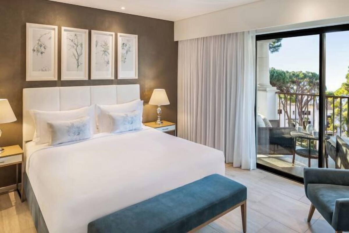 Une chambre d’hôtel avec un grand lit, une table de chevet, une lampe, un banc et un balcon aménagé avec du mobilier d’extérieur.