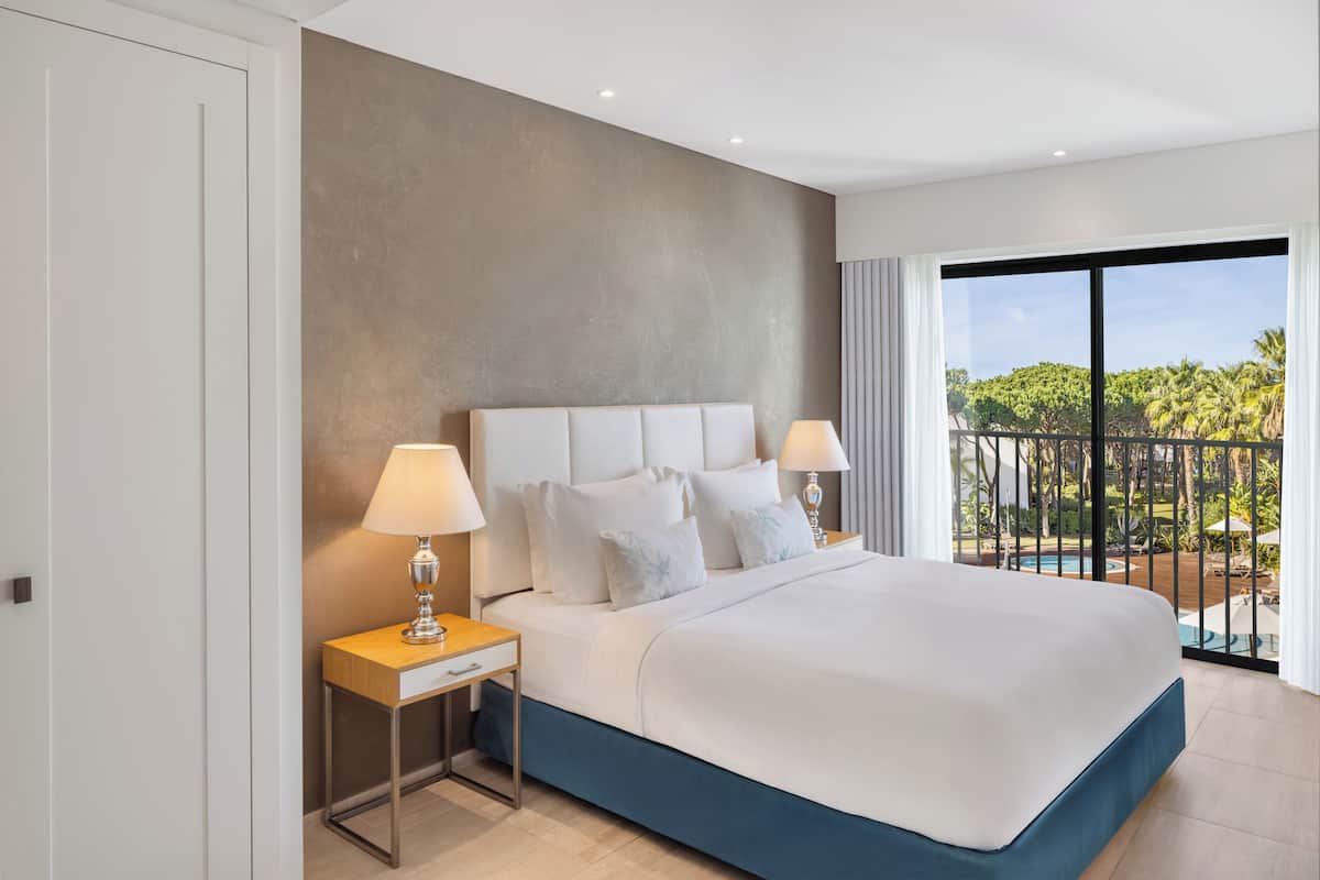 Une chambre d’hôtel moderne avec un grand lit, deux tables de chevet avec des lampes, et une vue sur une piscine et la verdure grâce à de grandes fenêtres.