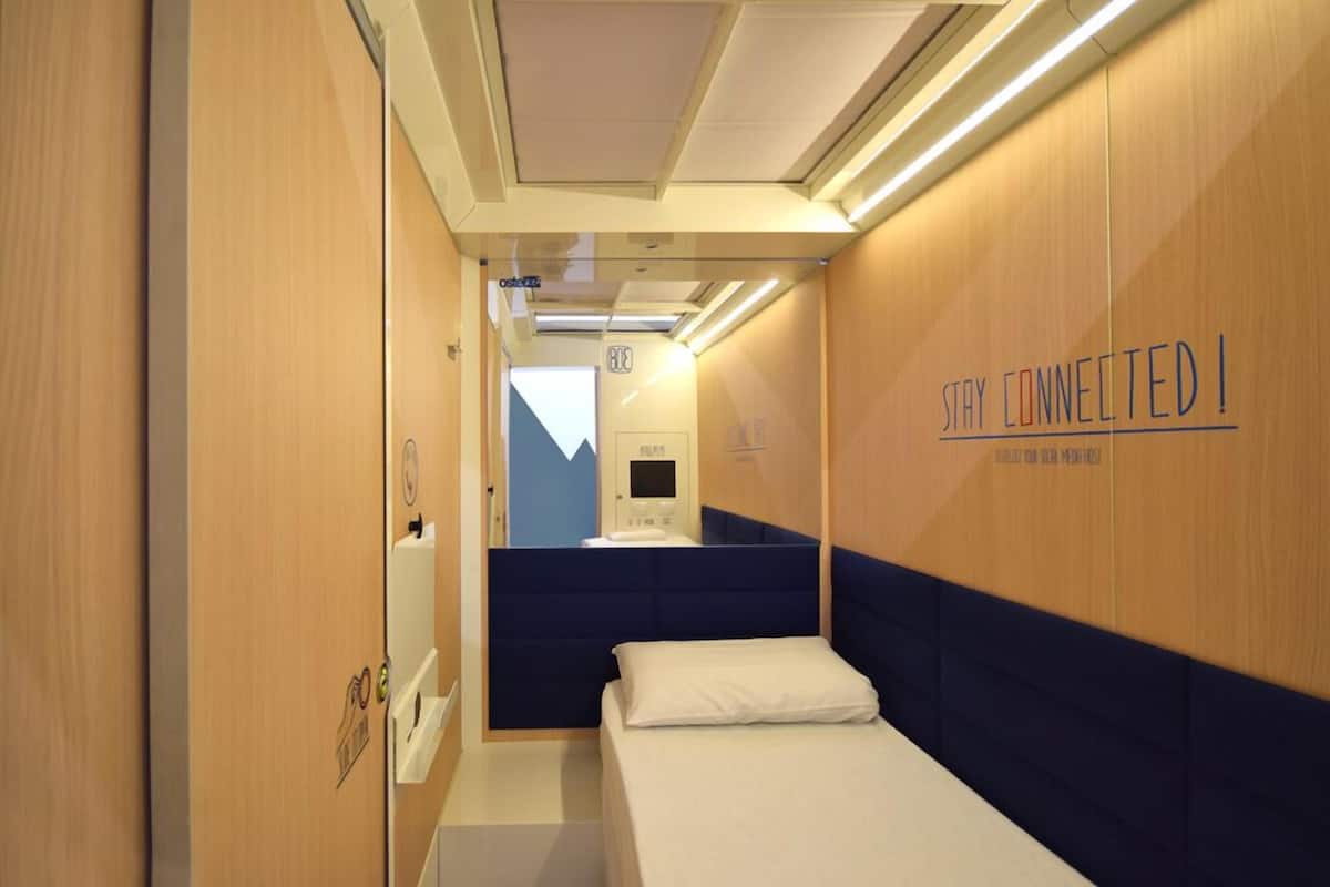 A narrow, modern train compartment with wooden walls, a single bed with a blue headboard, and a sign that reads "STAY CONNECTED!"