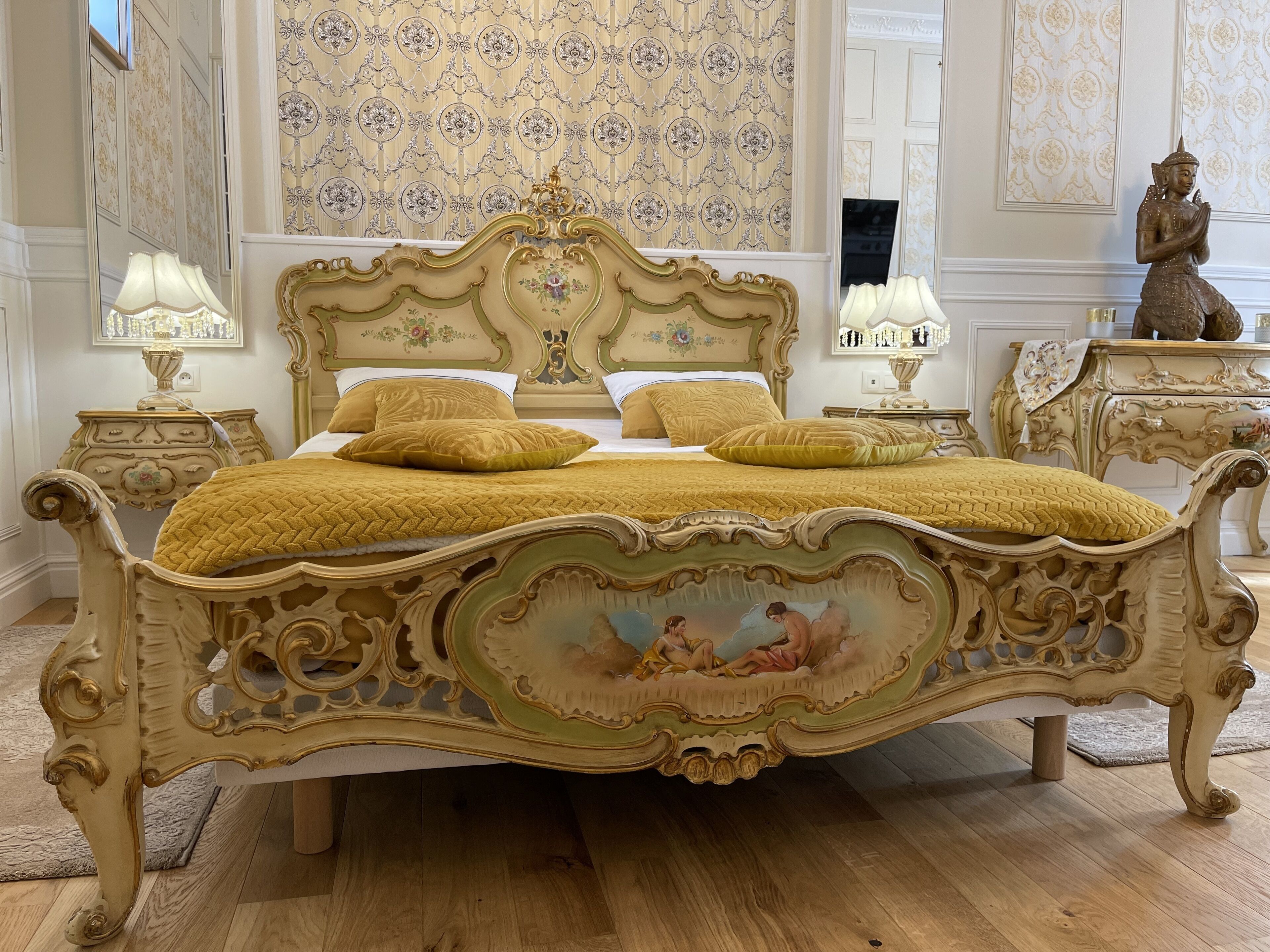 Ein klassisches, aufwendig gestaltetes Bett mit detailreichem Kopfteil und floralen Bildern, platziert in einem stilvoll eingerichteten Zimmer mit passender Möbel und gemustertem Wandpapier.