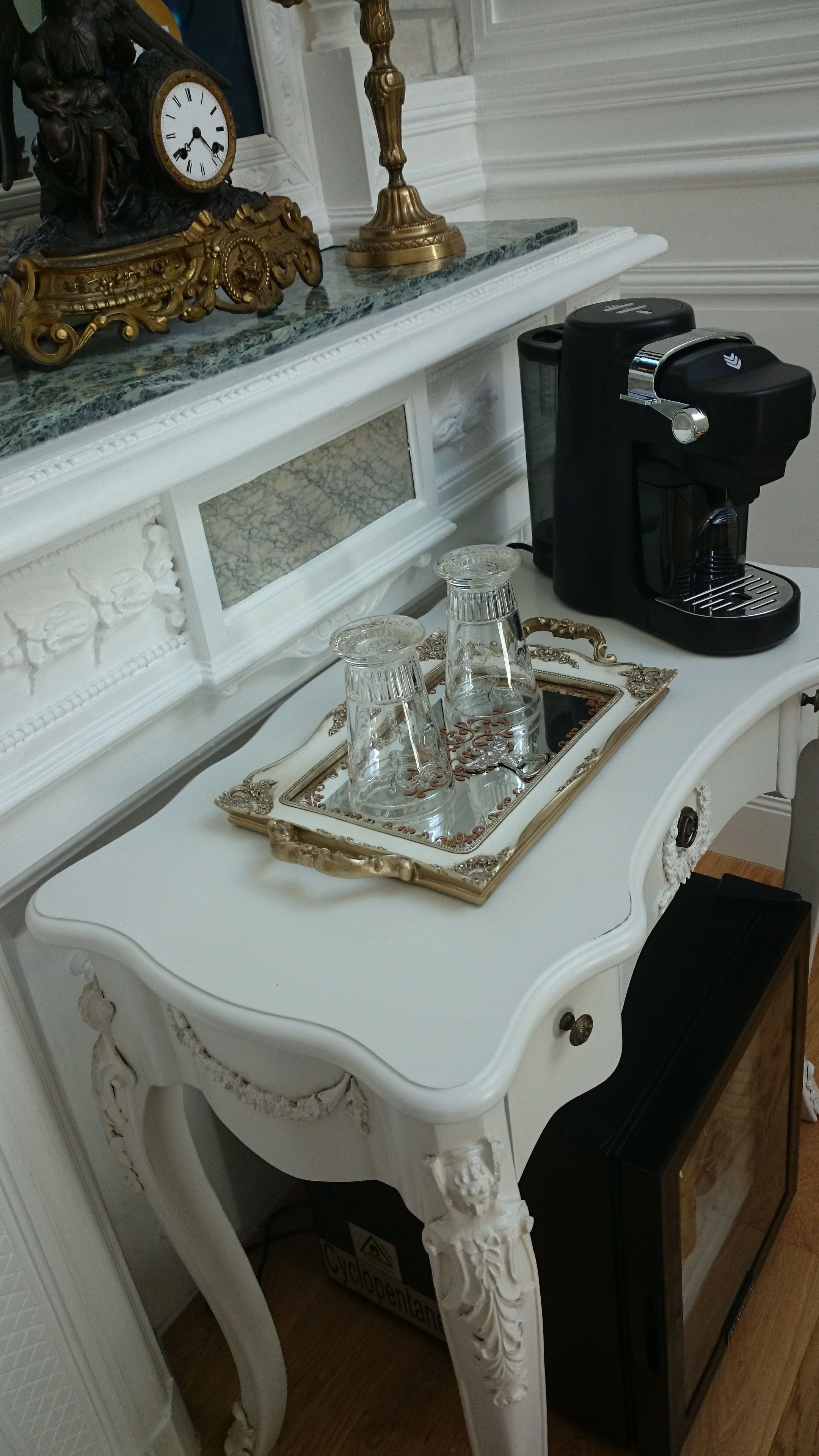 Een witte tafel met een dienblad waarop twee glazen staan en een decoratief object. Op de tafel staat een koffiezetapparaat, en erboven op de schoorsteenmantel hangt een klok.