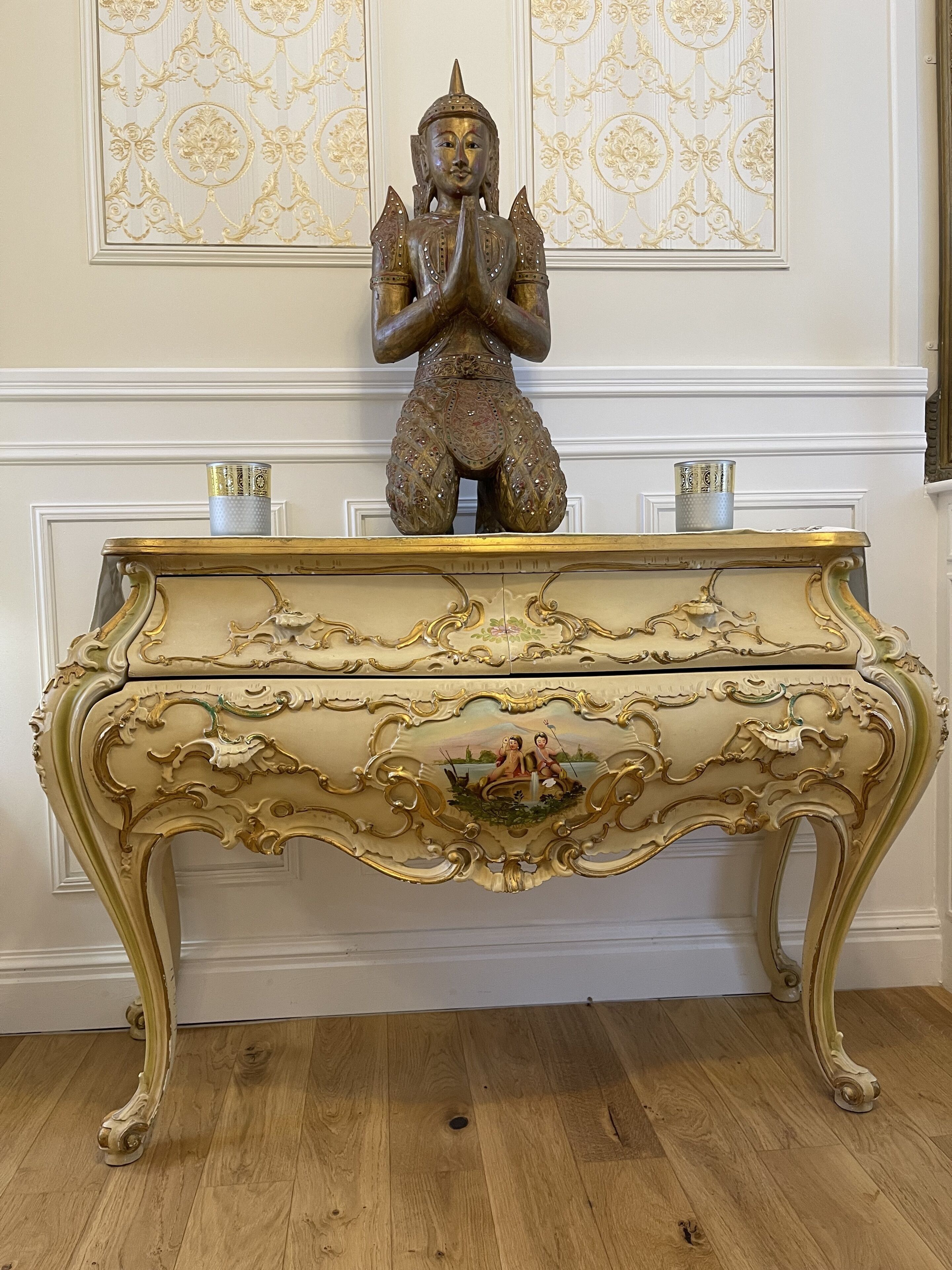 A statue of a seated figure with hands in a prayer position, placed on a decorative console table with intricate carvings and a central relief.