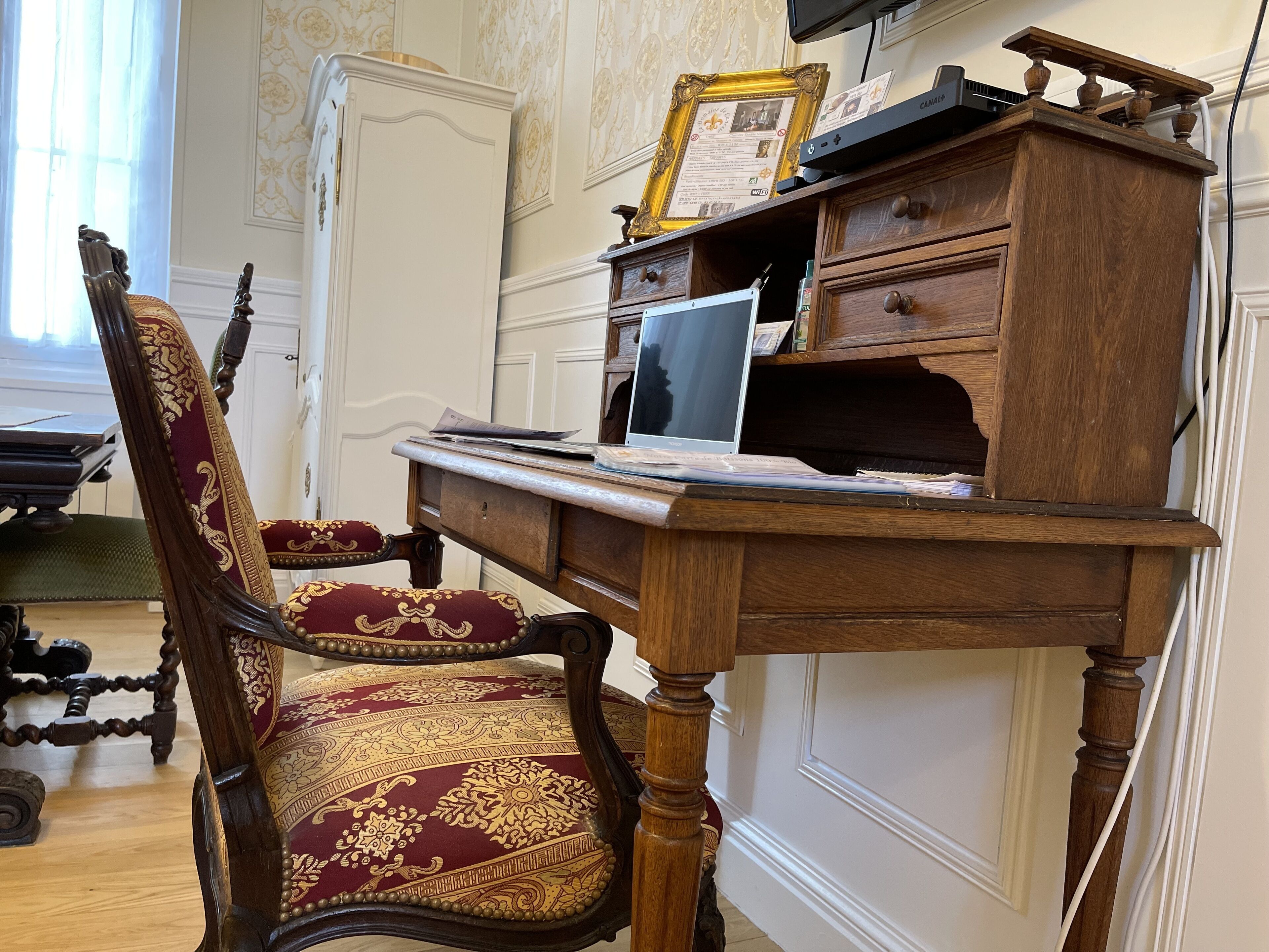A wooden desk with a laptop, a framed certificate, and a wooden chair with a red and gold patterned upholstery.