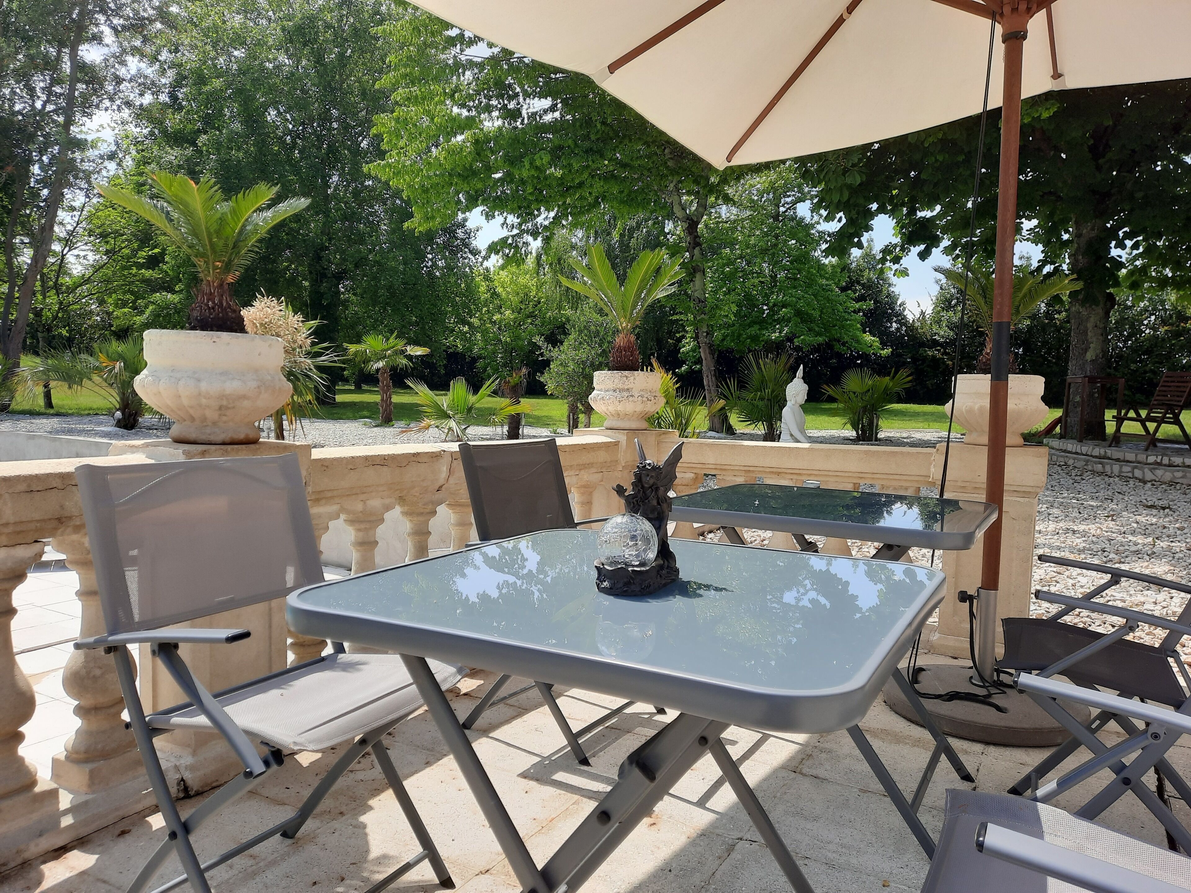Une terrasse extérieure avec une table et des chaises, entourée de plantes en pot et d’une statue.