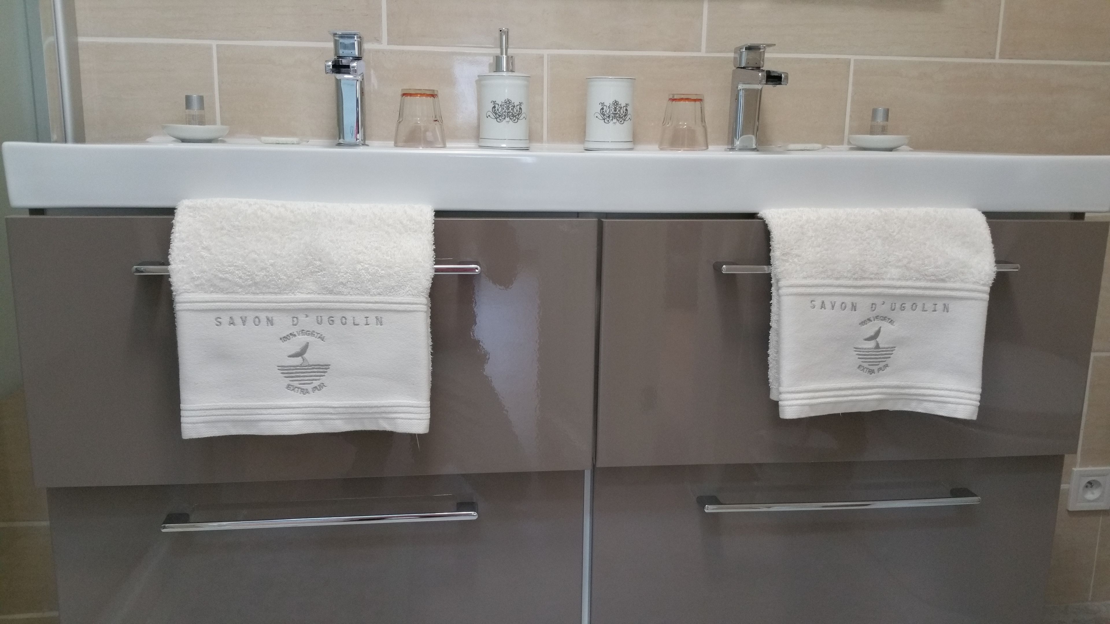 A bathroom with two sinks, each with a white towel hanging on a metal towel rack. Above the sinks, there are various toiletries and a soap dish.