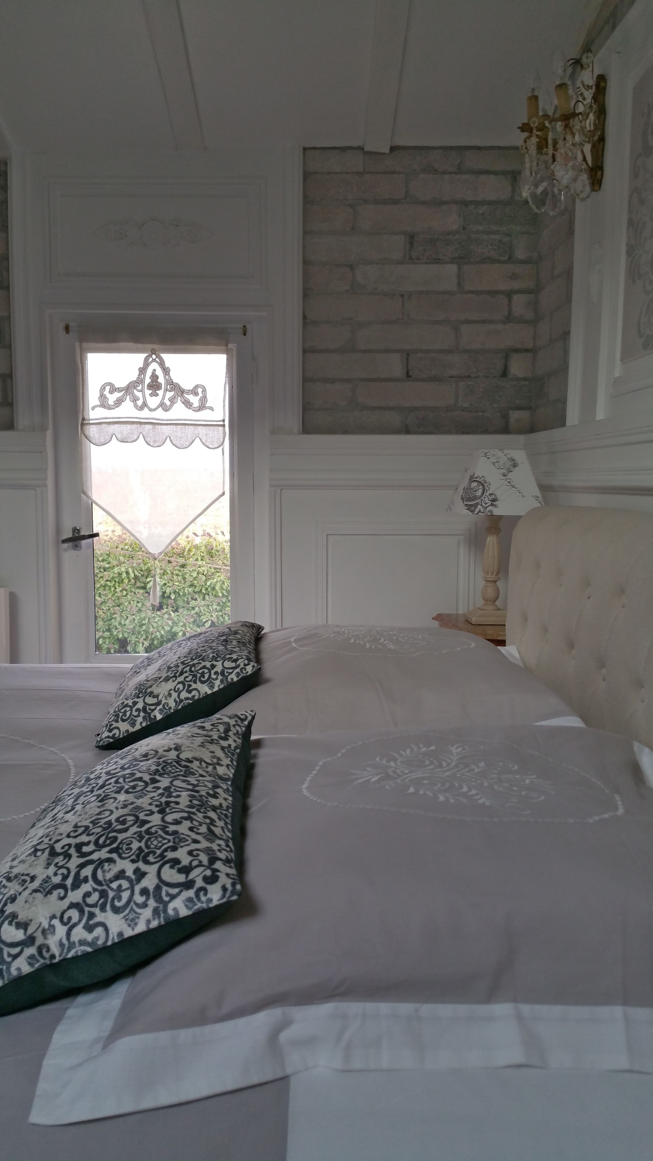 Une chambre à coucher avec un lit, des coussins à motifs, une tête de lit en bois, une table de chevet avec une lampe, une porte ornée d’un vitrail et un mur en briques.