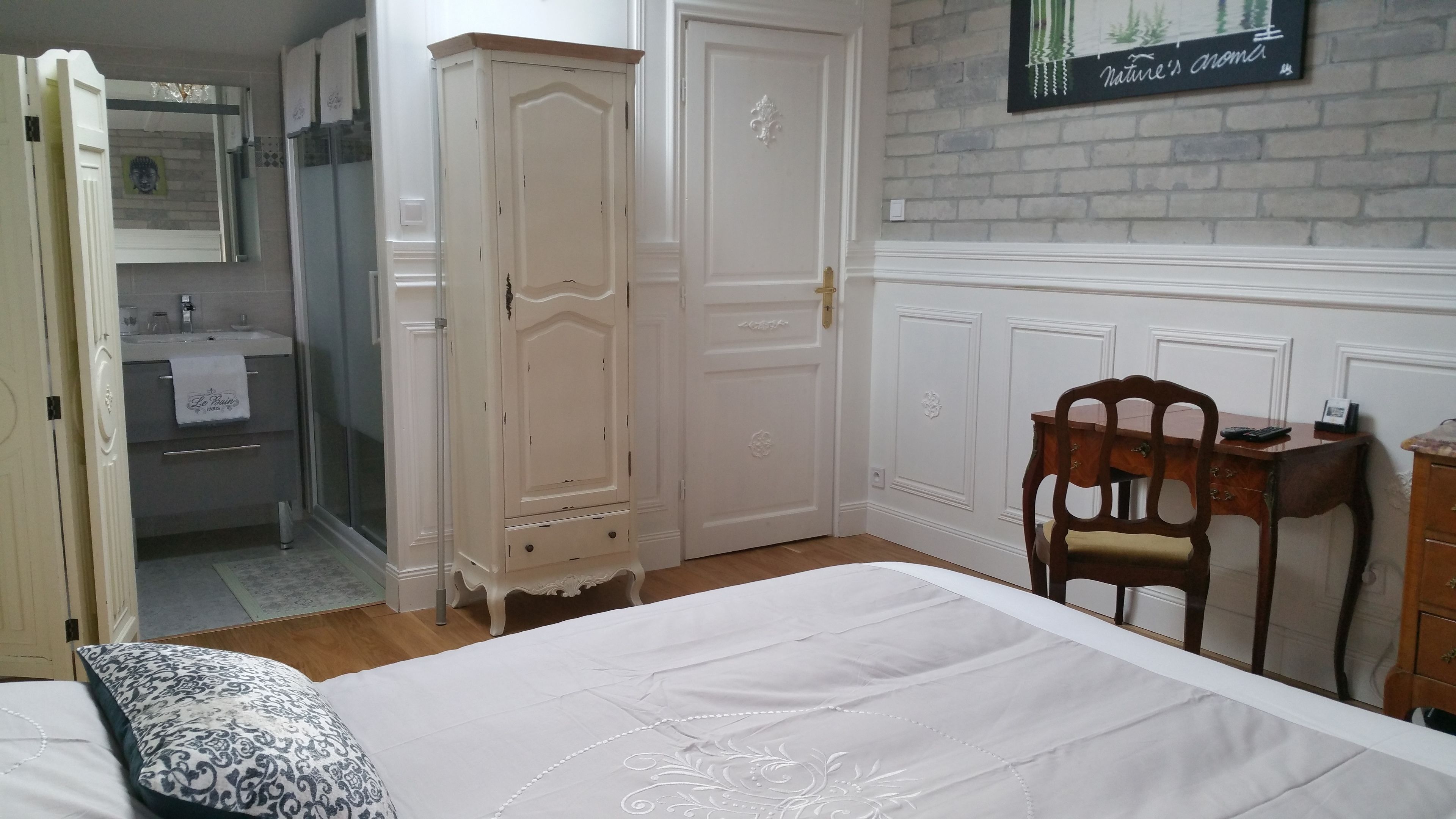A bedroom with a bed, a wooden chair, a desk with a chair, a wardrobe, a mirror, and a bathroom with a sink and towel.