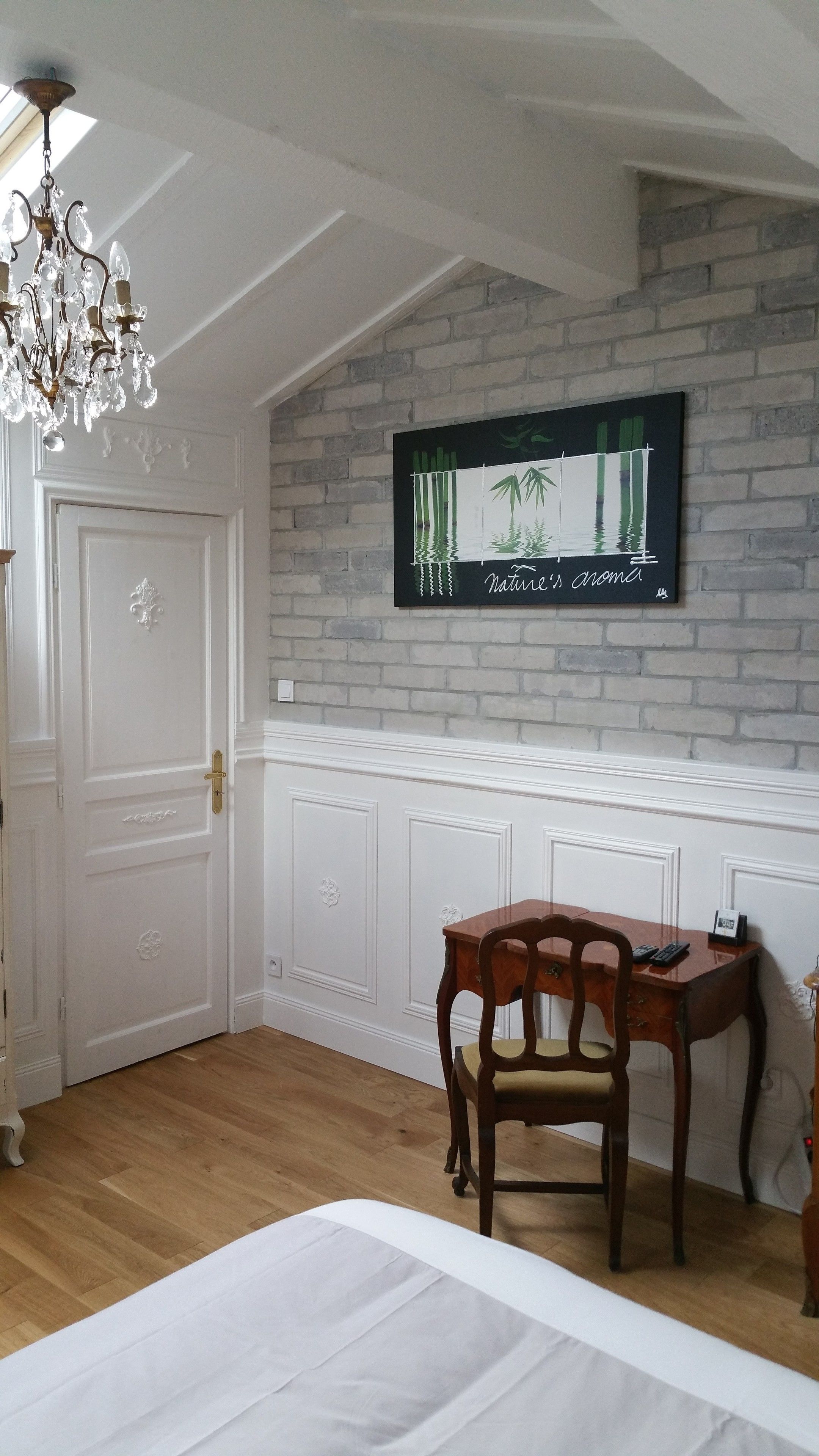Une pièce avec une porte blanche, un bureau en bois avec une chaise, un lustre et un tableau représentant des bambous accroché au mur.