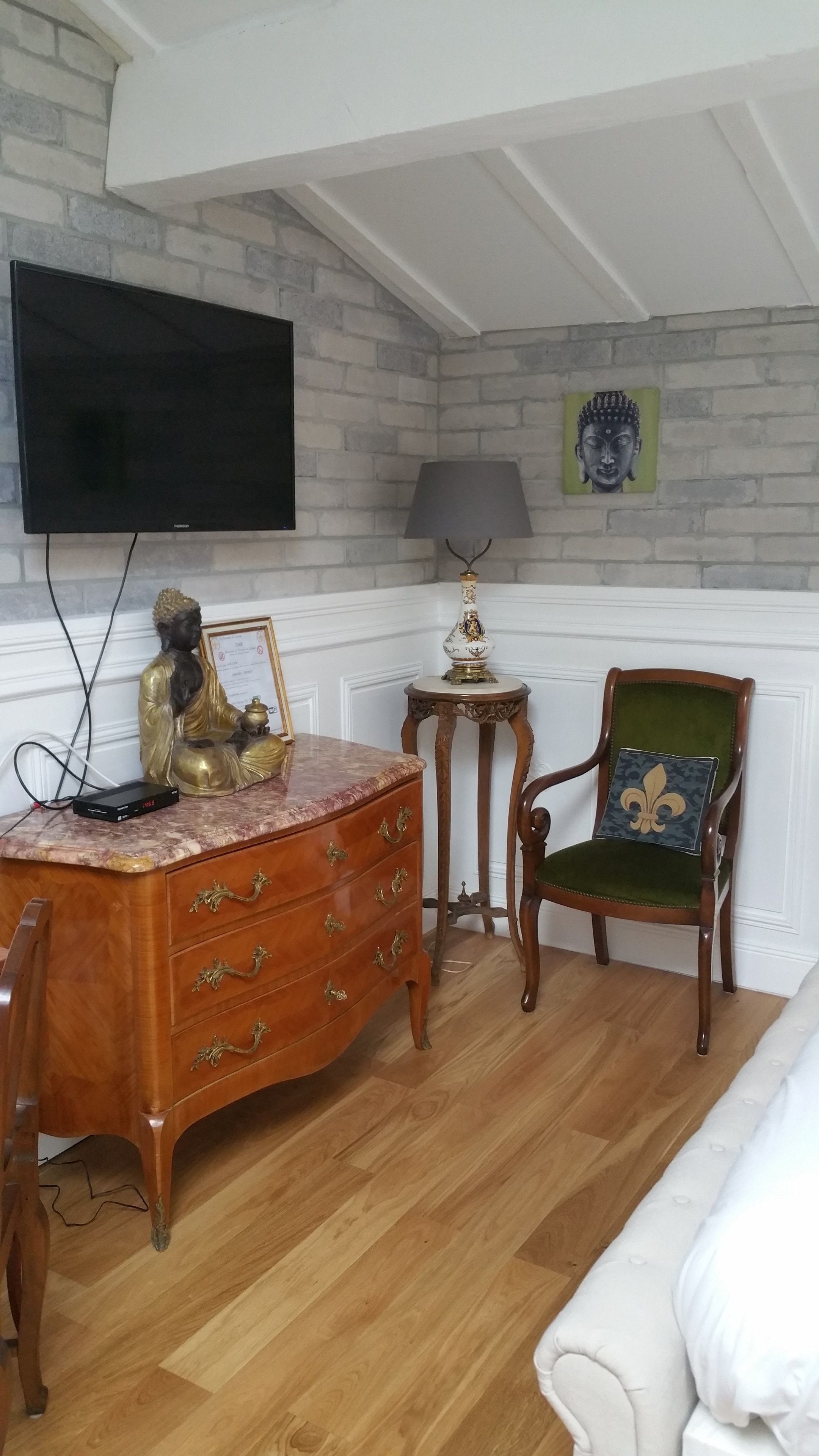 Una habitación con un escritorio de madera, un televisor en la pared, una estatua de Buda, una lámpara, una silla y una cama.