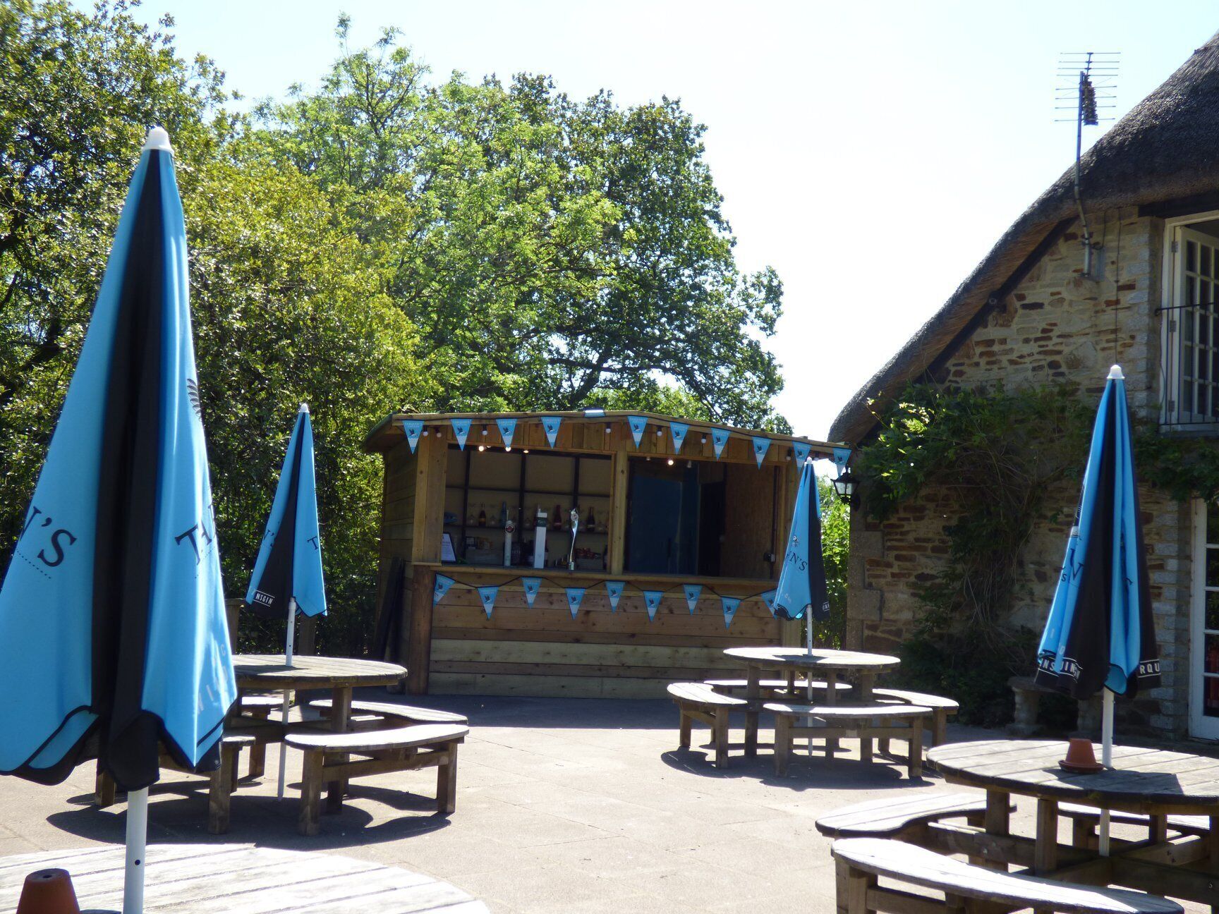 Un’area salotto esterna rustica con tavoli di legno e ombrelloni blu, una kiosk di legno con tetto di paglia e un edificio in pietra sullo sfondo.