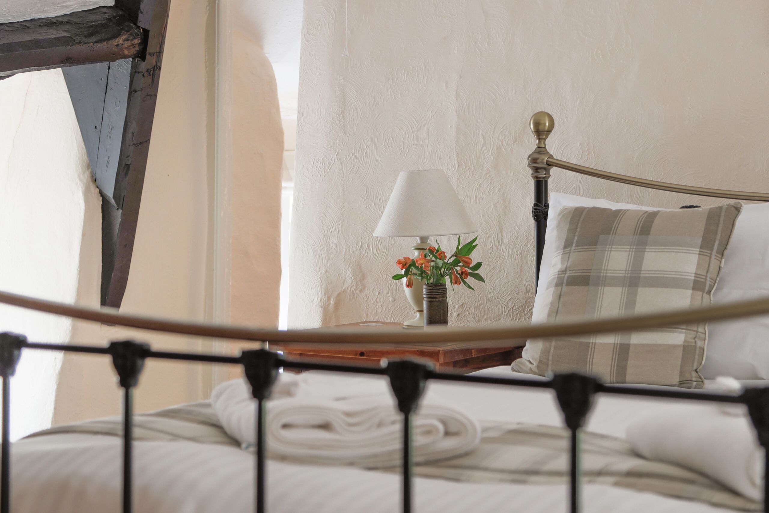 Ein ordentlich bezogenes Bett mit weißer Tagesdecke und Kissen in Karomuster, ein Nachttisch mit Lampe und Blumenvase sowie ein Fenster mit durchsichtigen Vorhängen.