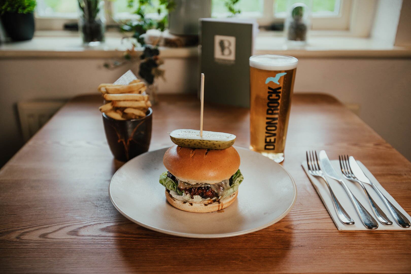 Una hamburguesa con pepinillo encima, una cerveza en un vaso, papas fritas y un menú sobre una mesa de madera junto a una ventana.