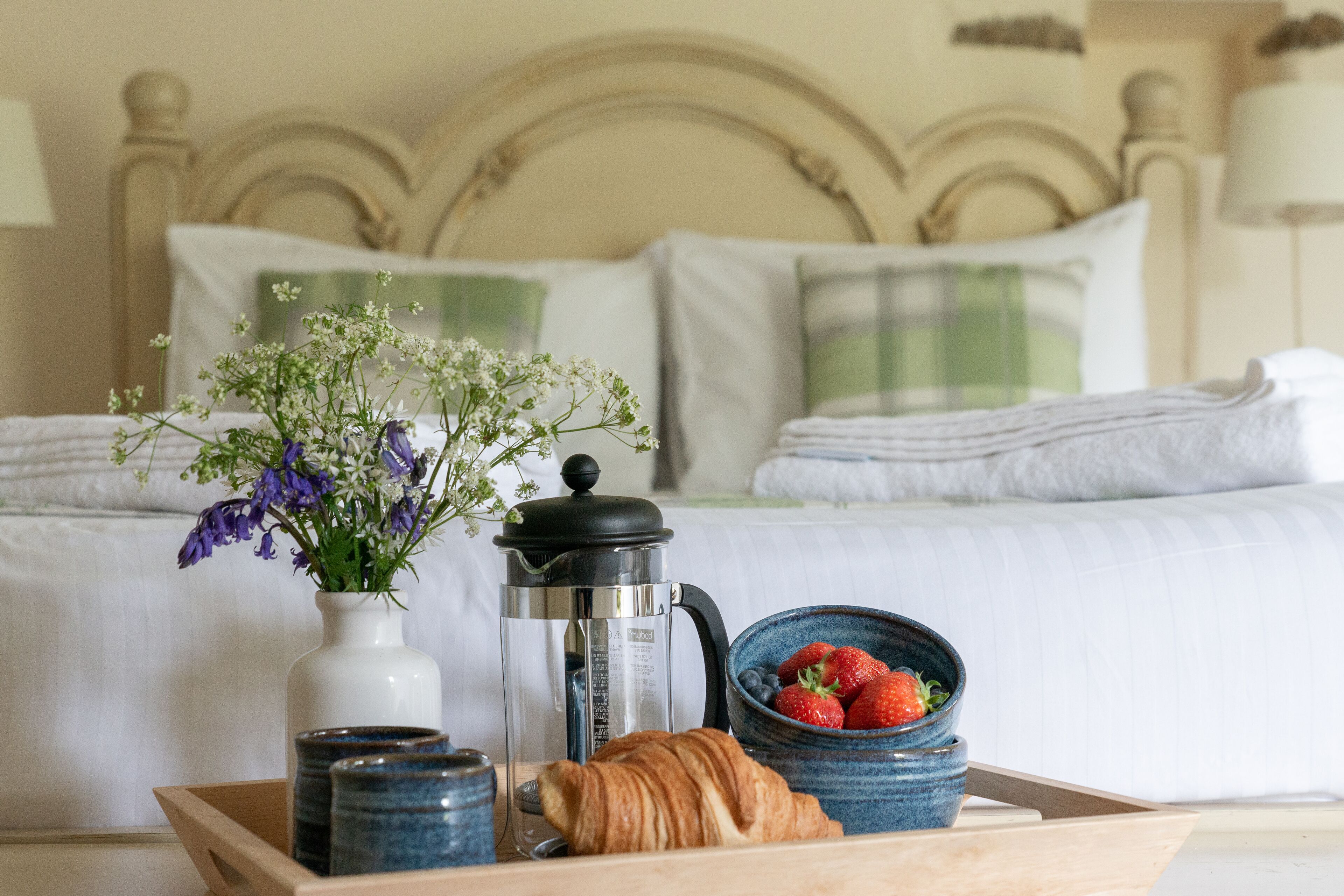 En bakke med morgenmad, bestående af en croissant, en skål jordbær, en kaffepotte og en vase med blomster, på et træbord i et hotelværelse.
