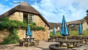 A stone building with a thatched roof, surrounded by wooden picnic tables and blue umbrellas. - Bearslake Inn (Okehampton)
