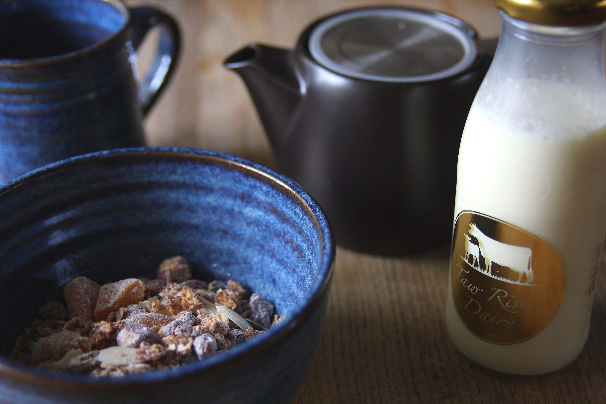 Eine Schüssel mit Frühstücksflocken, ein Teekessel und eine Milchflasche auf einer Holztischplatte.