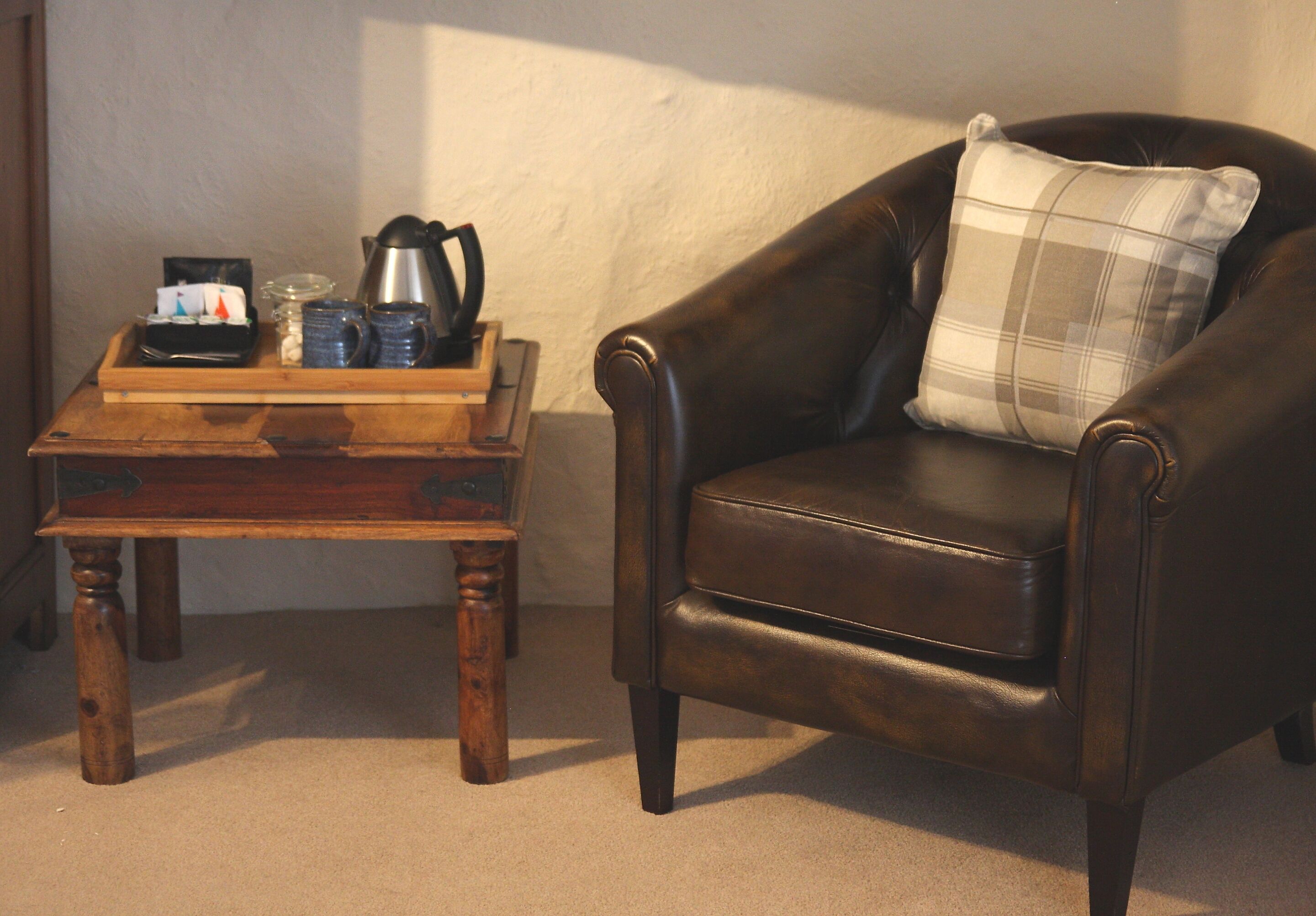 A dark brown leather armchair with a plaid pillow, a wooden coffee table with a tray of items, and a wall-mounted shelf.