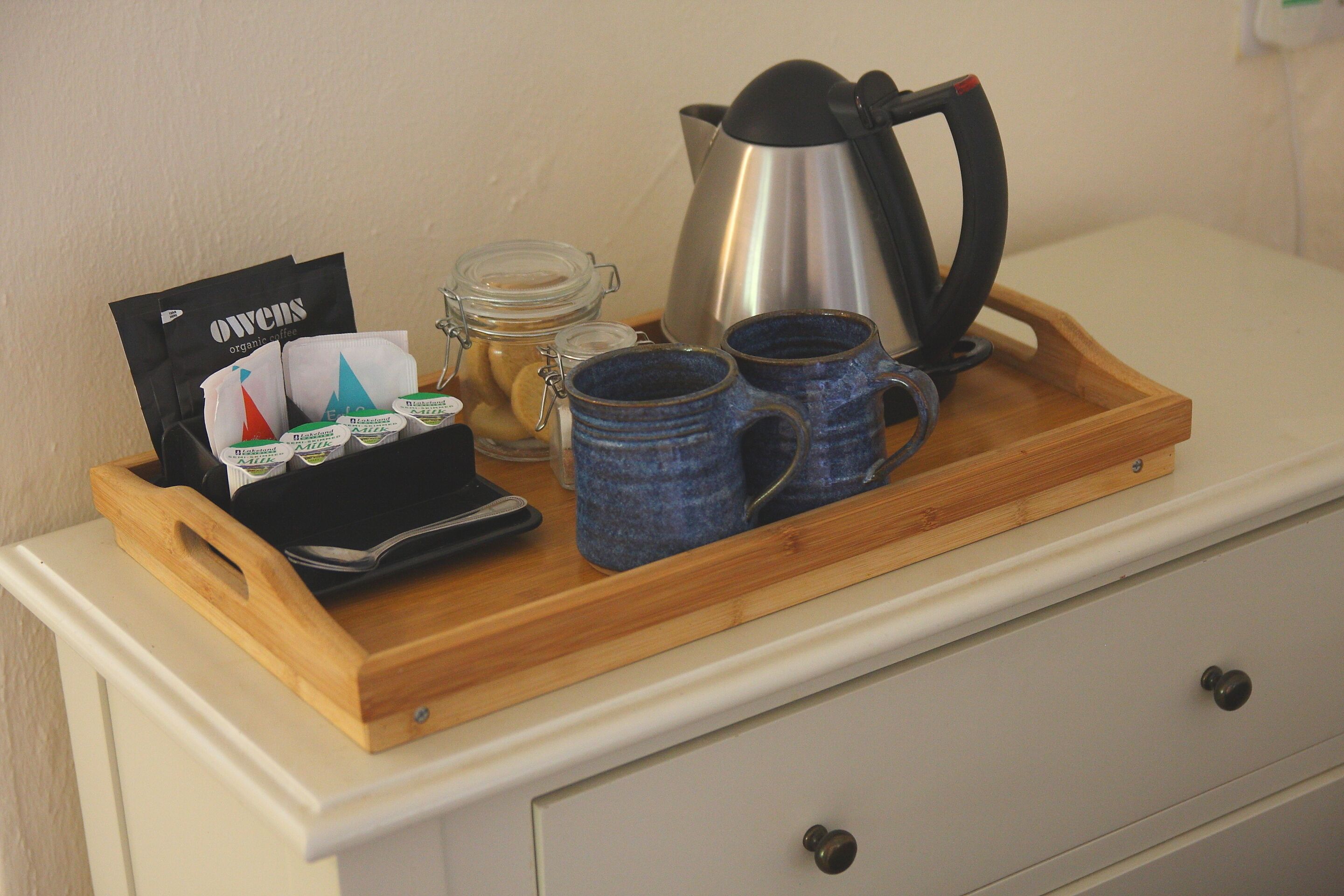 A tray with a coffee maker, mugs, a jar of jam, and a bag of coffee pods on a white dresser.