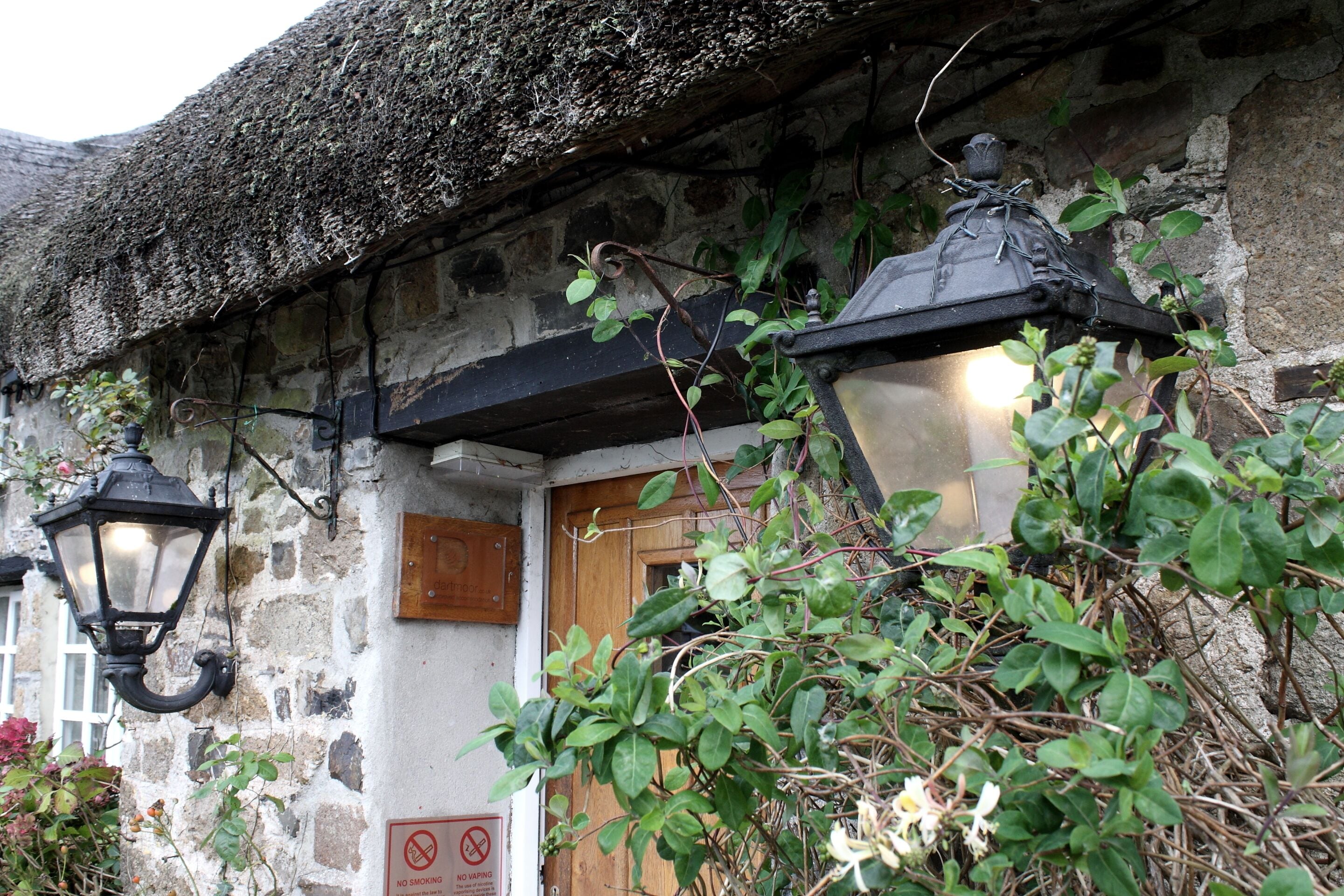 Un bâtiment rustique en pierre, avec un toit de chaume, une porte en bois et deux lampes de style lanterne, de couleur noire.