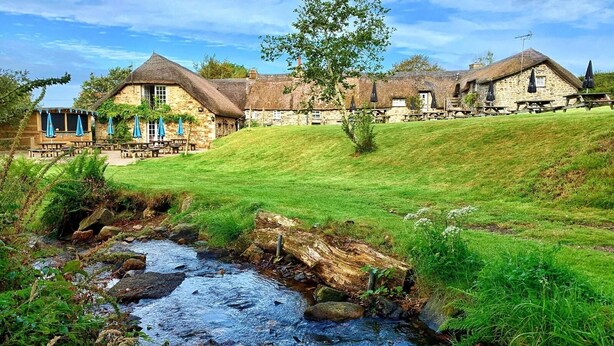 A stone building with a thatched roof, a grassy hill, and a stream. - Bearslake Inn (Okehampton)