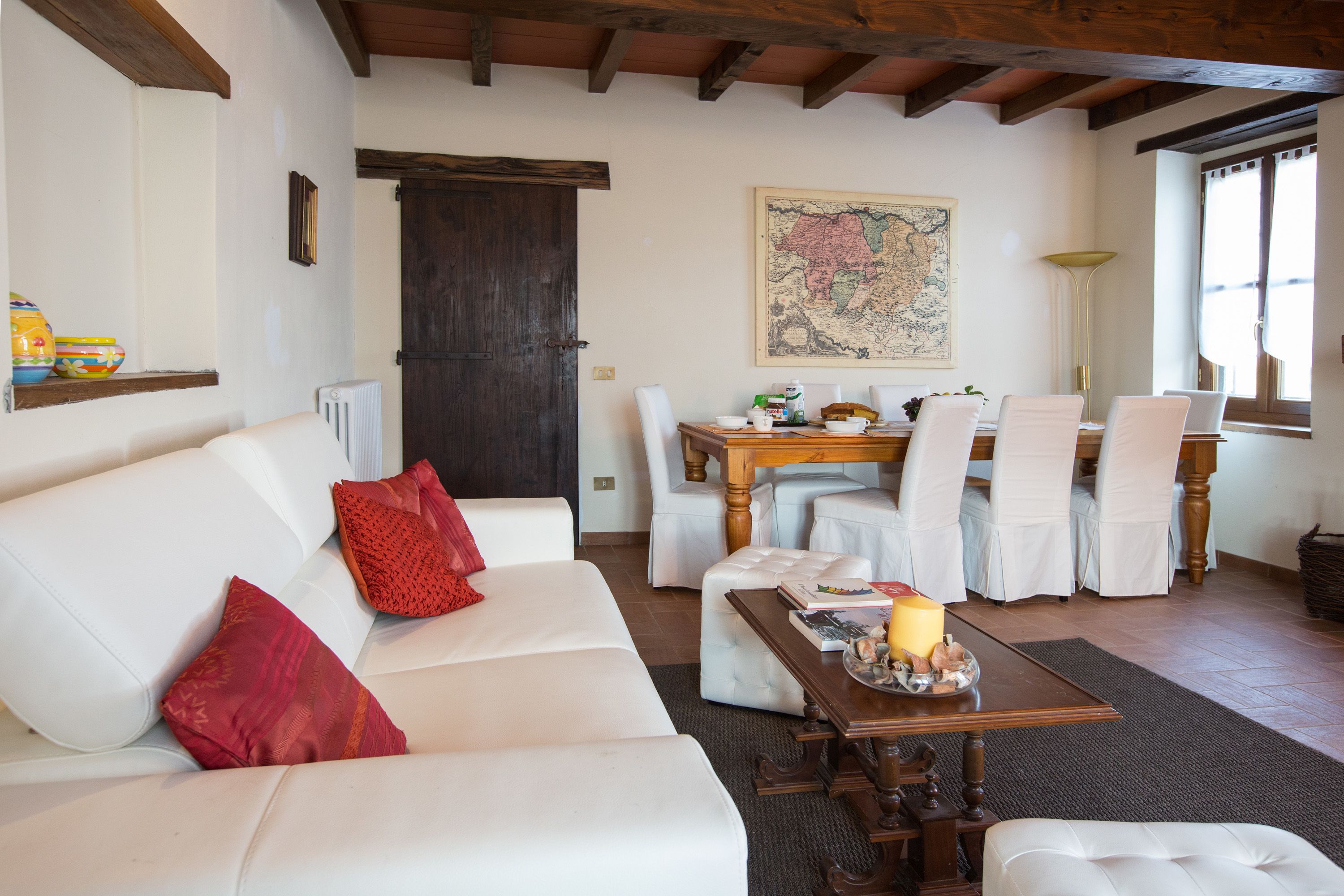 A cozy living room with a white sofa, red pillows, a wooden coffee table with a candle, and a dining area with white chairs and a wooden table.