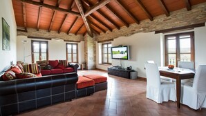 Ein großzügiger Wohnbereich mit hoher Holzbalkendecke, einem großen Flachbildfernseher, einem Esstisch mit Stühlen und einem Sofa mit roten Kissen.