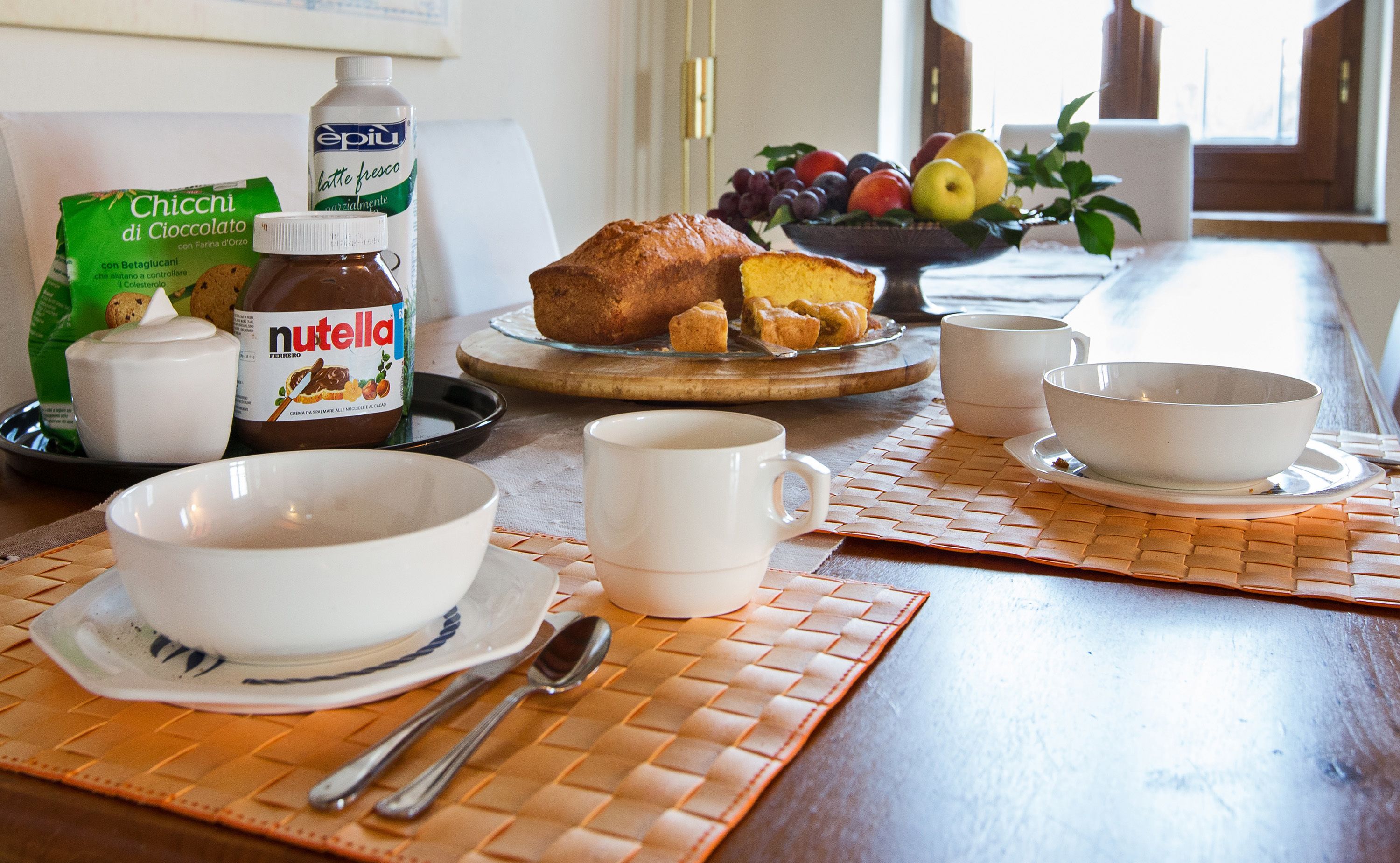 Un desayuno con hogaza de pan, un cuenco de fruta, un tarro de Nutella, un cartón de leche y una taza de café sobre una mesa.
