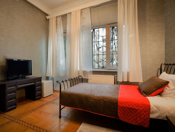 Un dormitorio con cama, televisor sobre un aparador y ventana con cortinas.