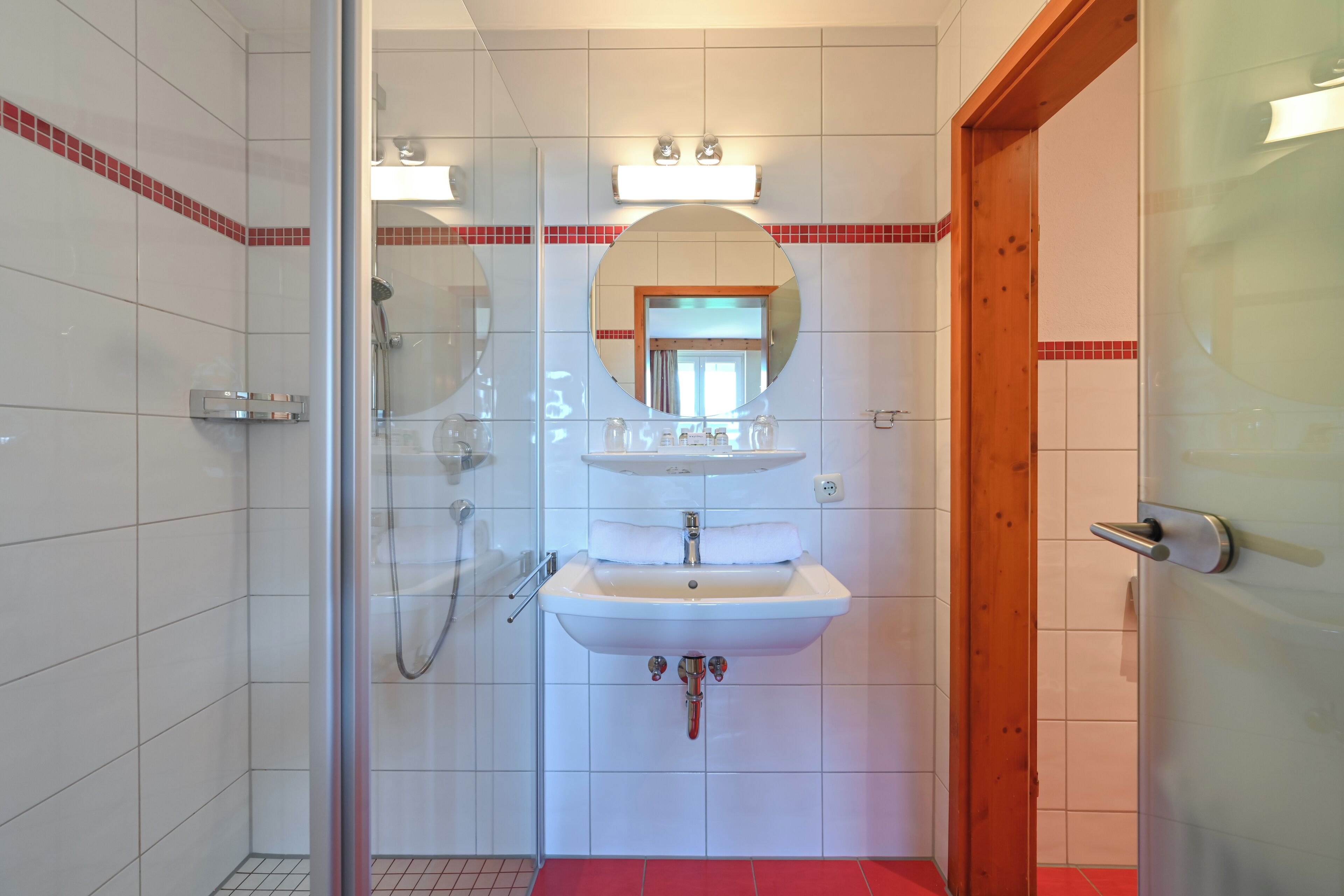 A bathroom with a glass shower enclosure, a white sink with a mirror above it, and a wooden door.