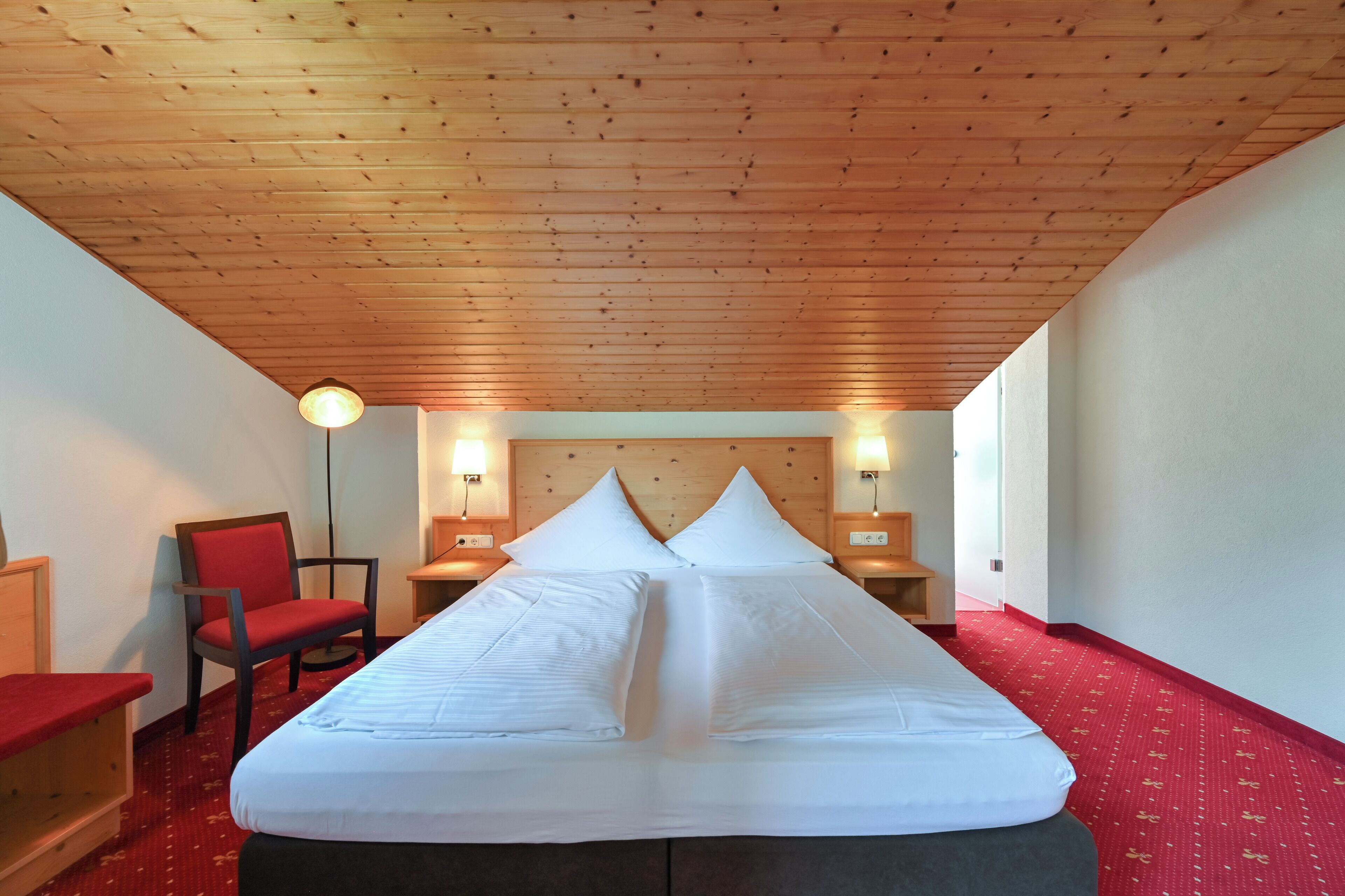 Ein Hotelzimmer mit Holzbalkendecke, einem großen Bett mit weißer Bettwäsche, einem roten Sessel und zwei Nachttischlampen.