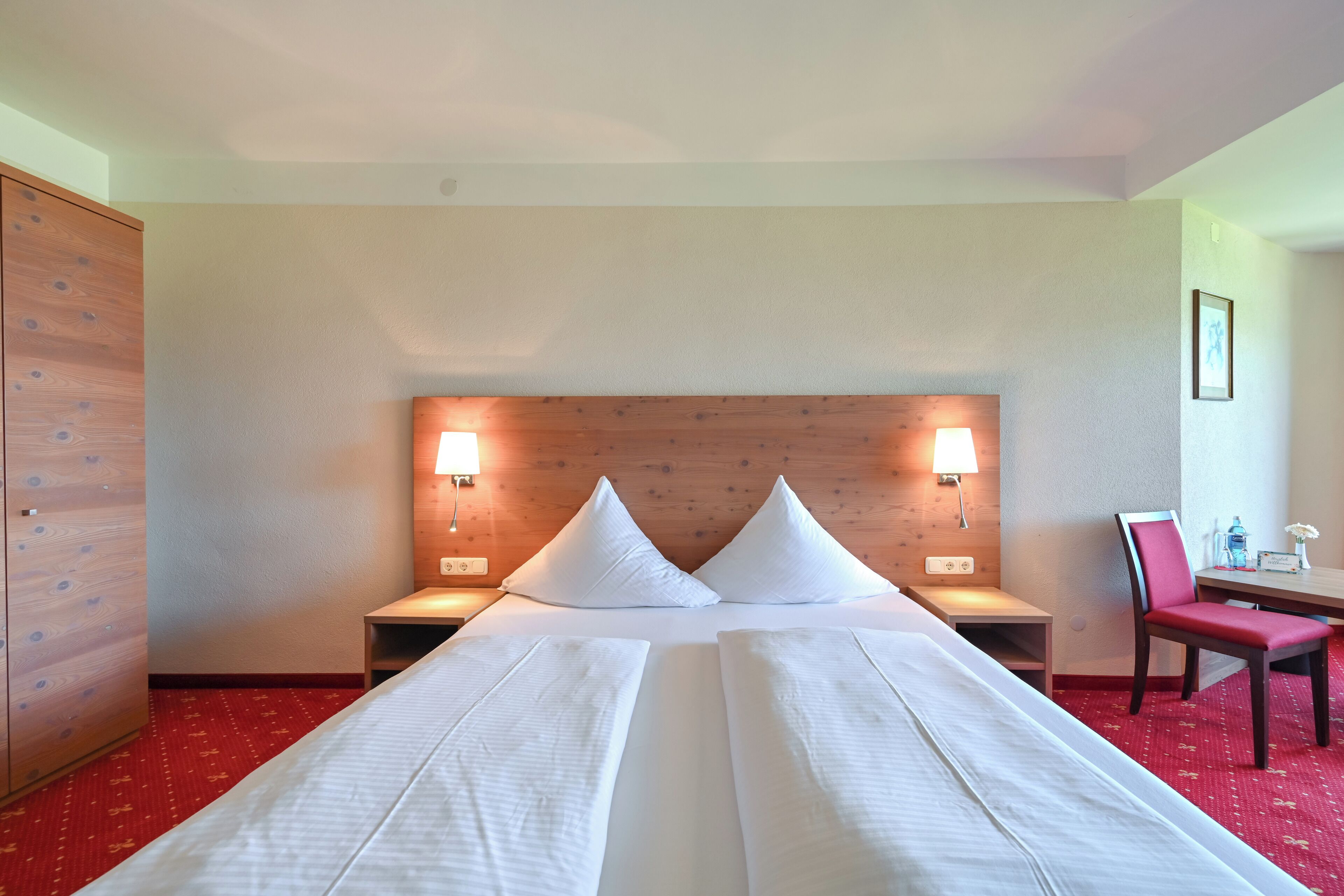 Quarto de hotel com cama de casal, duas mesas de cabeceira, cabeceira de madeira, tapete vermelho, guarda-roupa de madeira, escrivaninha com cadeira e duas luminárias fixadas na parede.