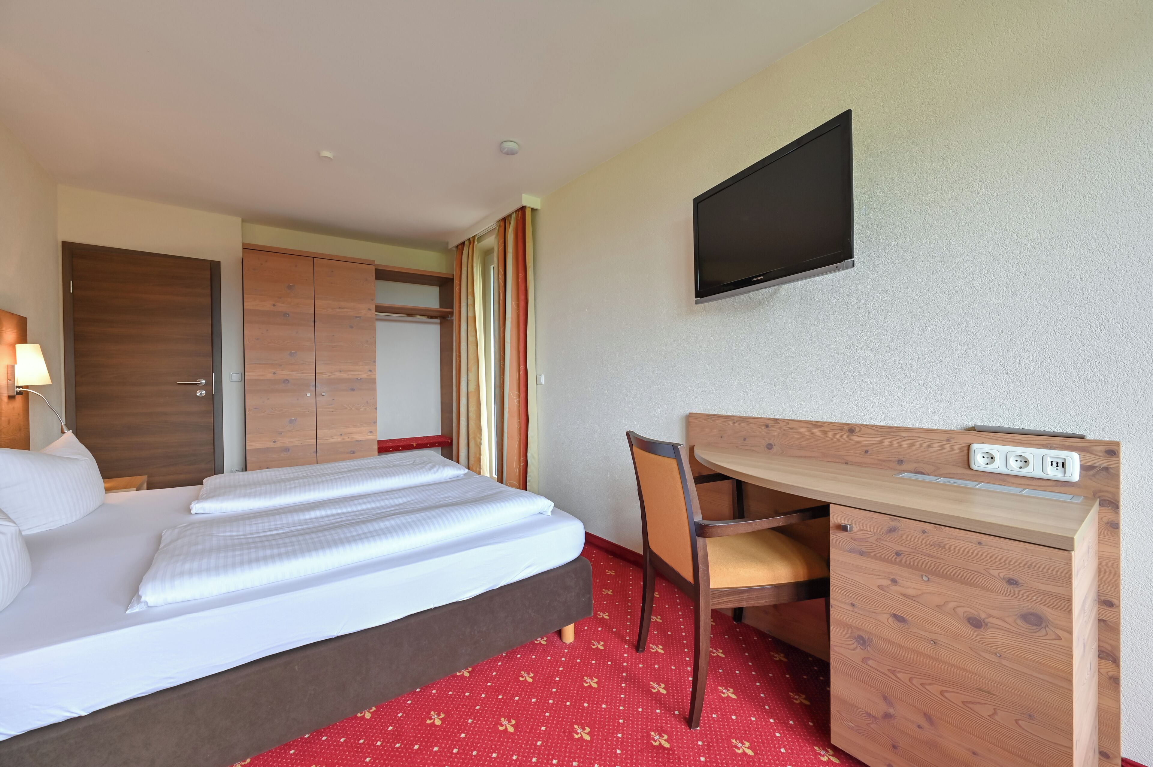 Habitación de hotel con cama, escritorio con silla, televisor montado en la pared y armario.