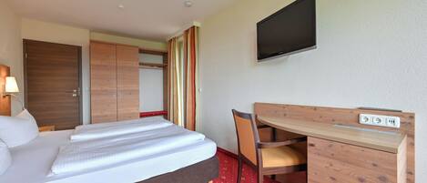 A hotel room with a bed, a desk with a chair, a TV mounted on the wall, and a wardrobe.