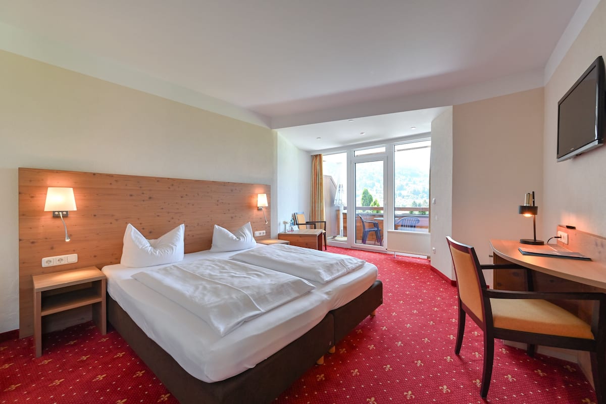 A hotel room with a large bed, a desk, and a chair. There is a window with a view of the outside.