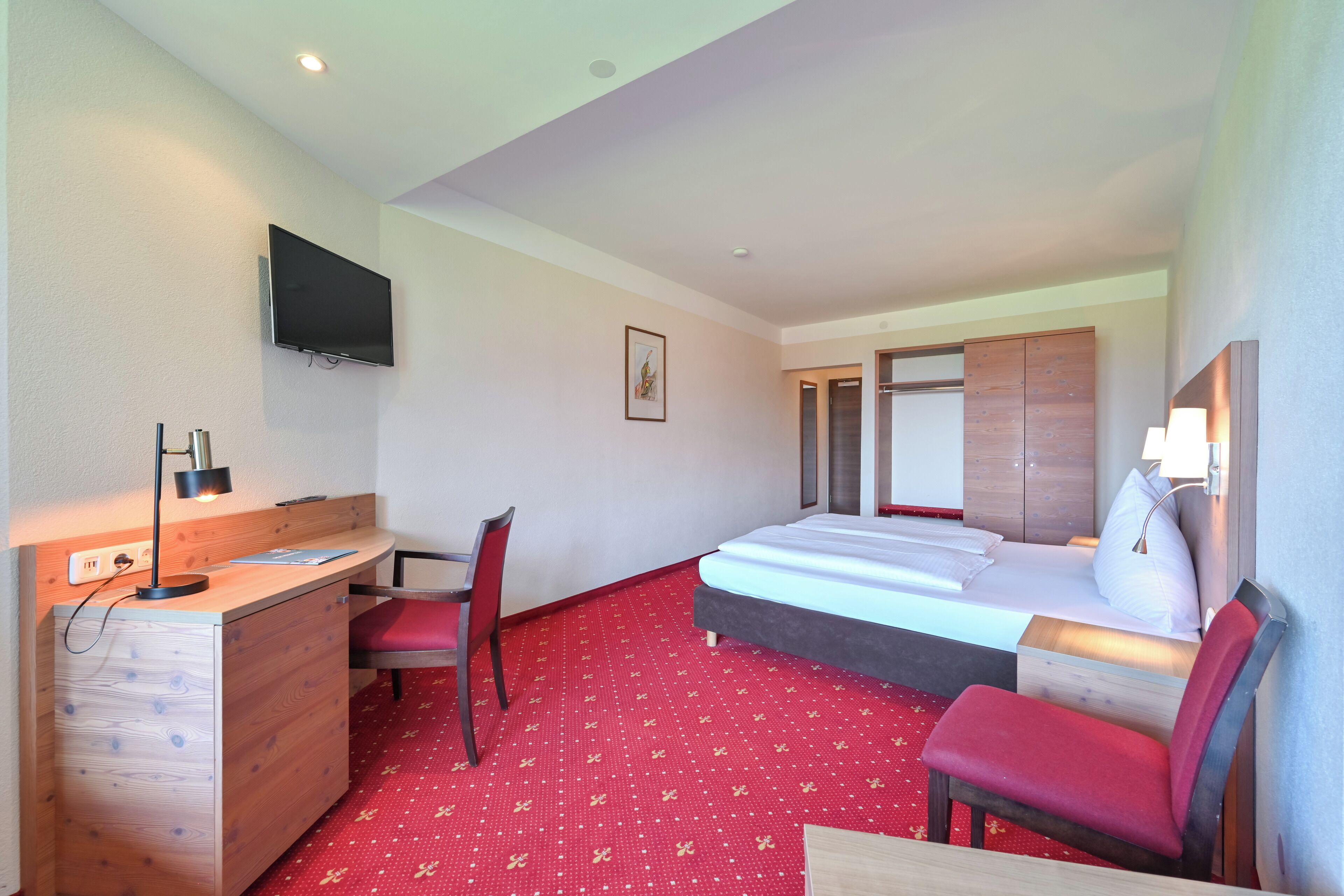A hotel room with a bed, a desk, a chair, a TV mounted on the wall, and a red carpeted floor.