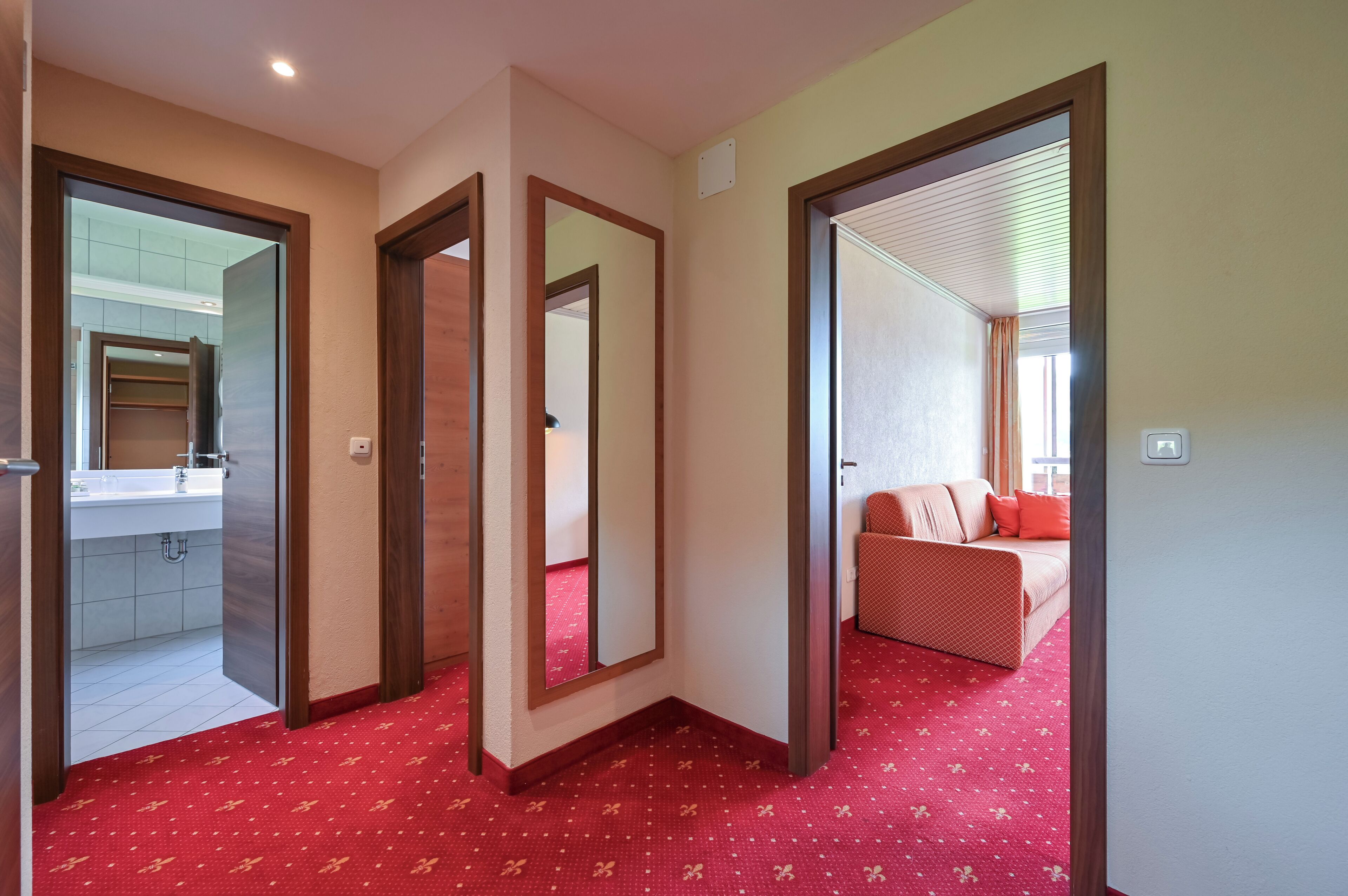 A hotel corridor with a red carpet, a mirror, and a view into a bedroom with a red sofa.