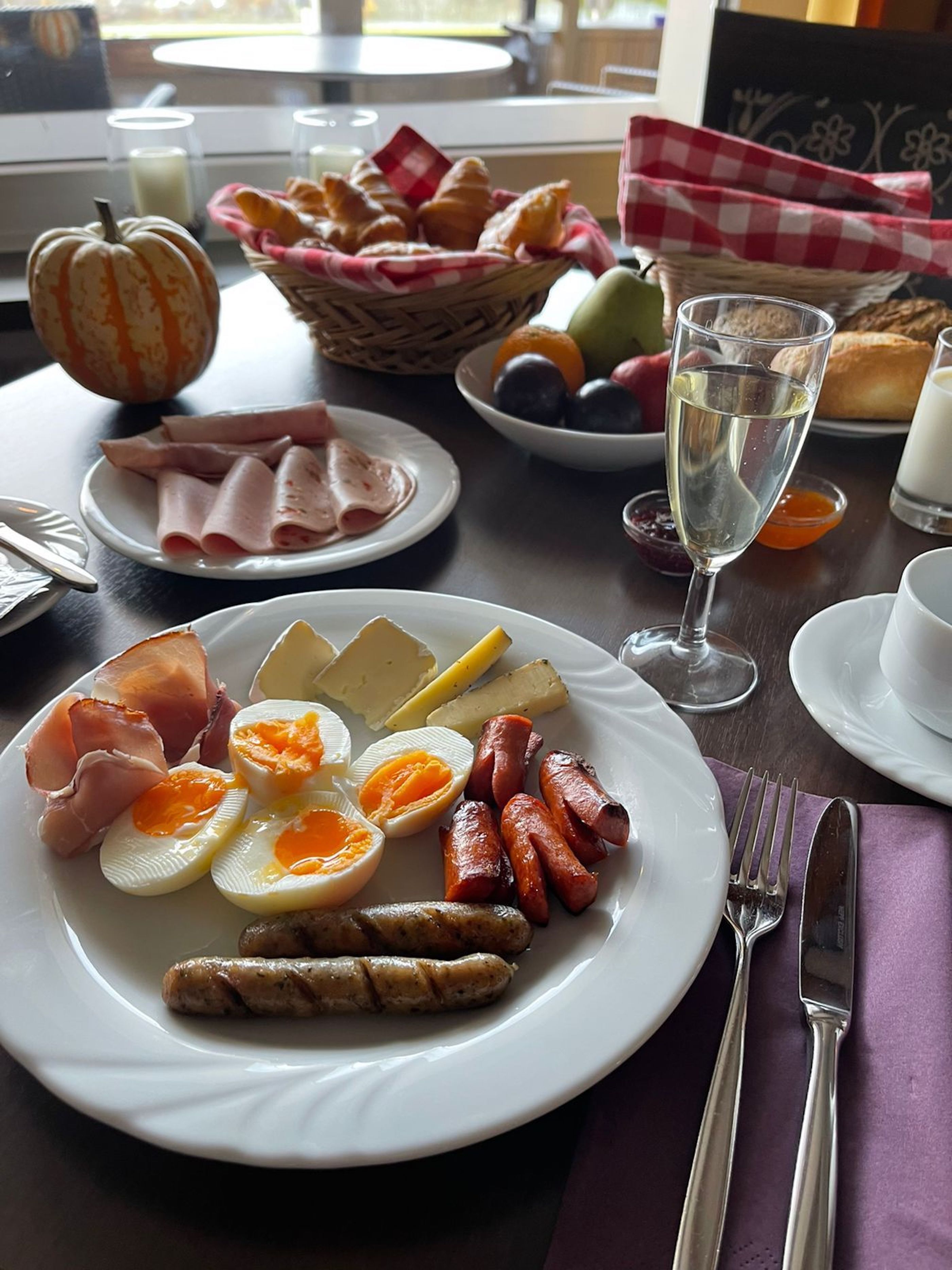 Un desayuno con un plato de huevos, jamón, salchichas y queso, una copa de vino blanco y una cesta de croissants.