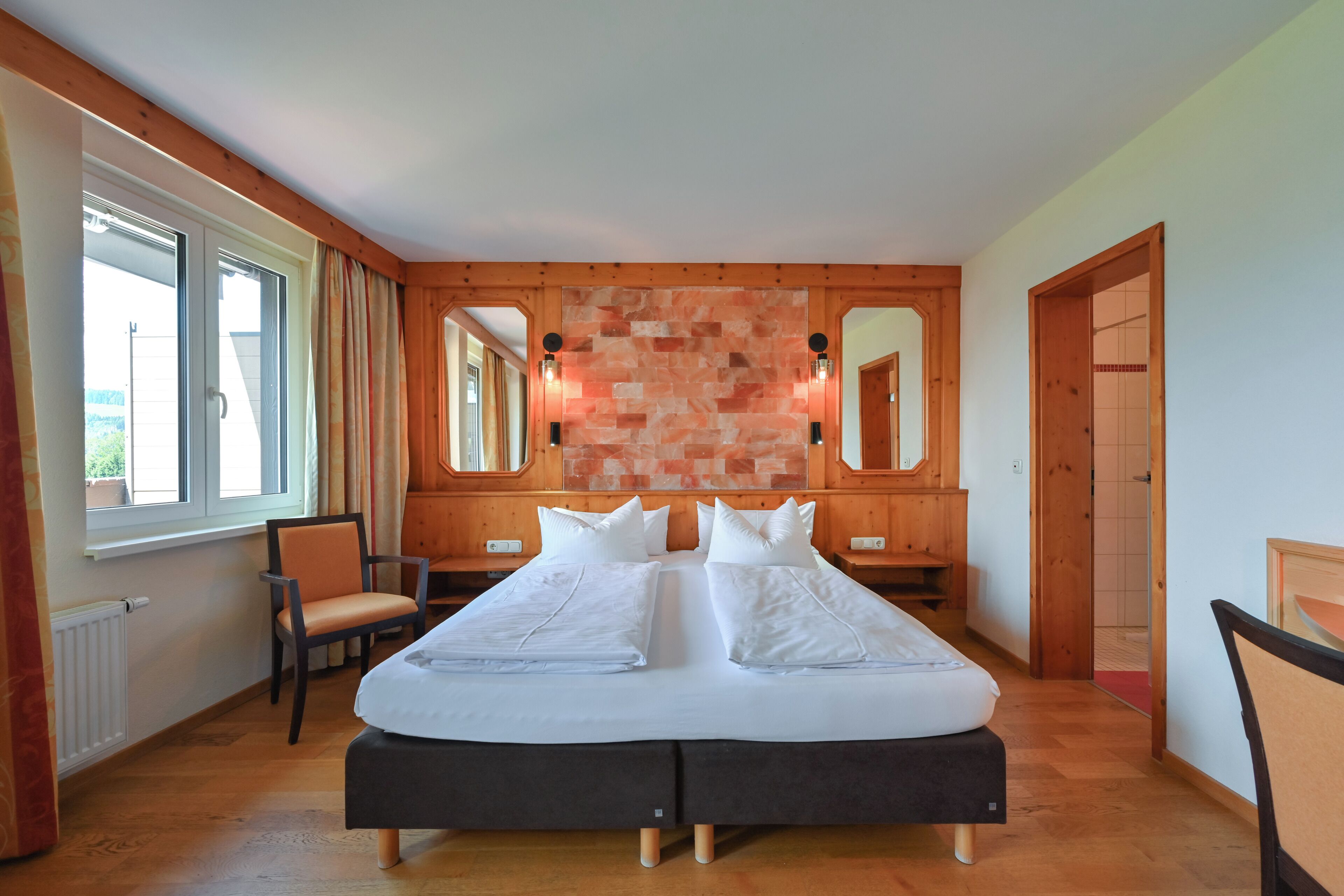 Una cama doble con cabecera oscura, paredes de madera y una pared de acento de piedra. Hay dos mesitas de noche con lámparas, una silla y una ventana con cortinas.