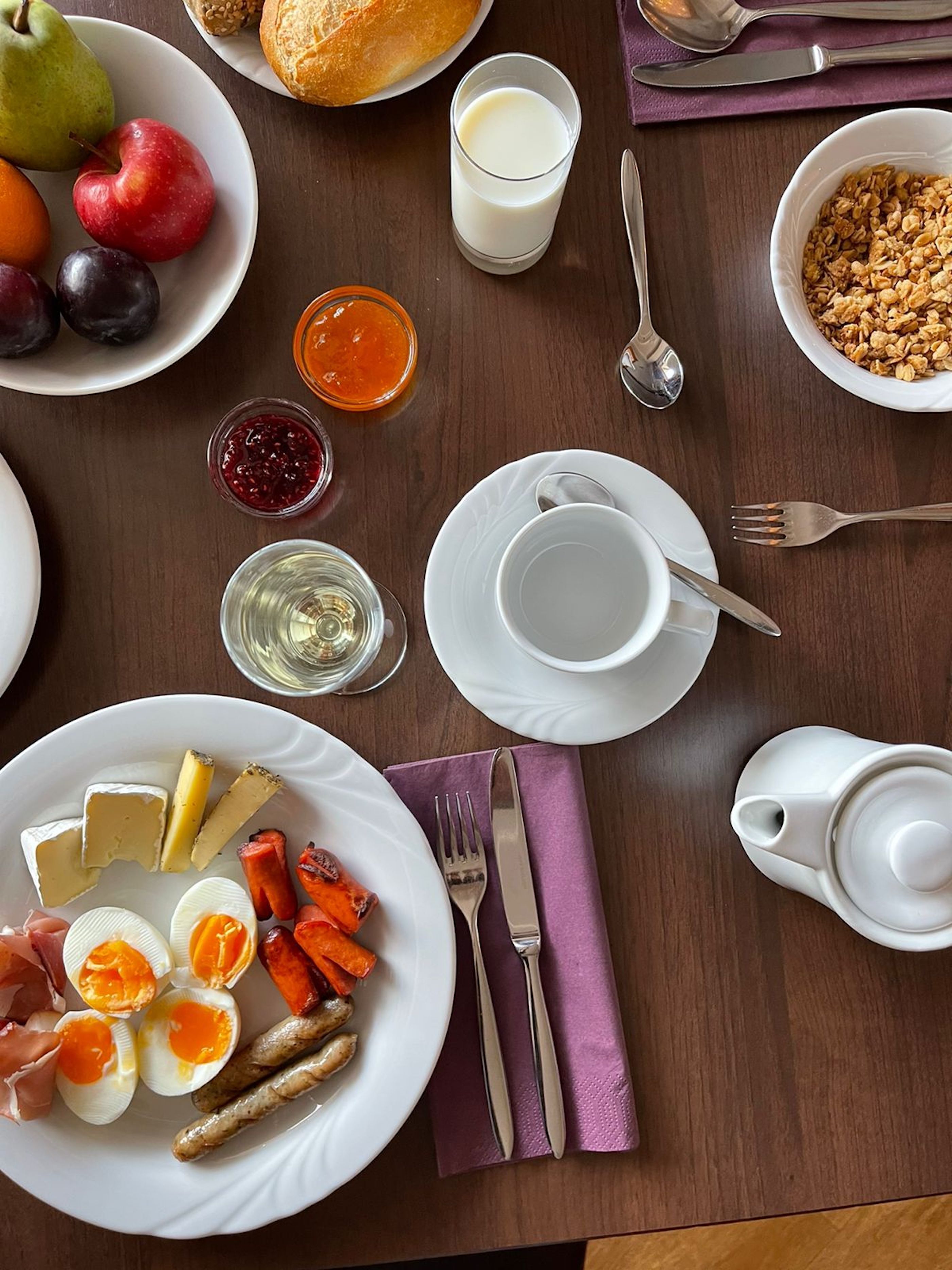 A breakfast setting with plates of eggs, cheese, and vegetables, a glass of milk, and a bowl of granola.