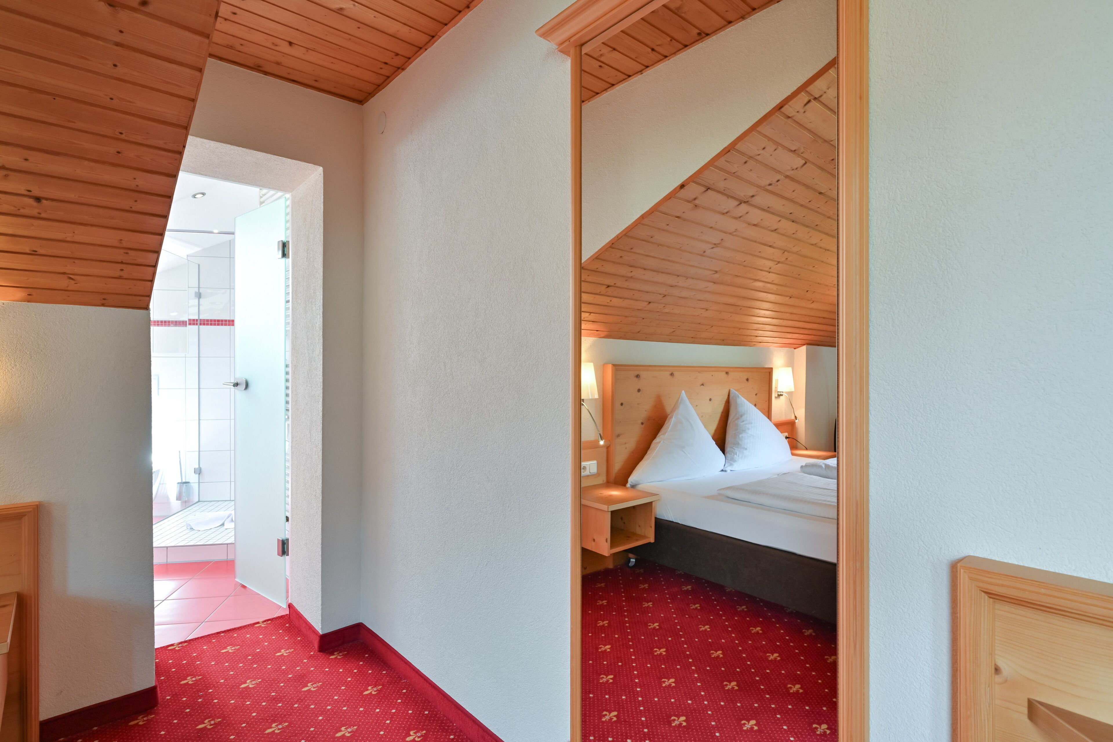 Habitación de hotel con techo de madera, cama con ropa blanca, mesita de noche y suelo cubierto de alfombra roja.
