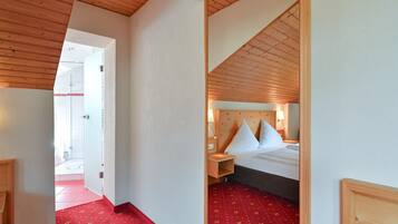 Habitación de hotel con techo de madera, cama con ropa blanca, mesita de noche y suelo cubierto de alfombra roja.