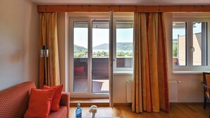 A room with a red sofa, a table with a vase and a bottle, and a view of a balcony with a wooden deck and mountains in the background. - Rothenfels Panorama Hotel (Immenstadt im Allgaeu)