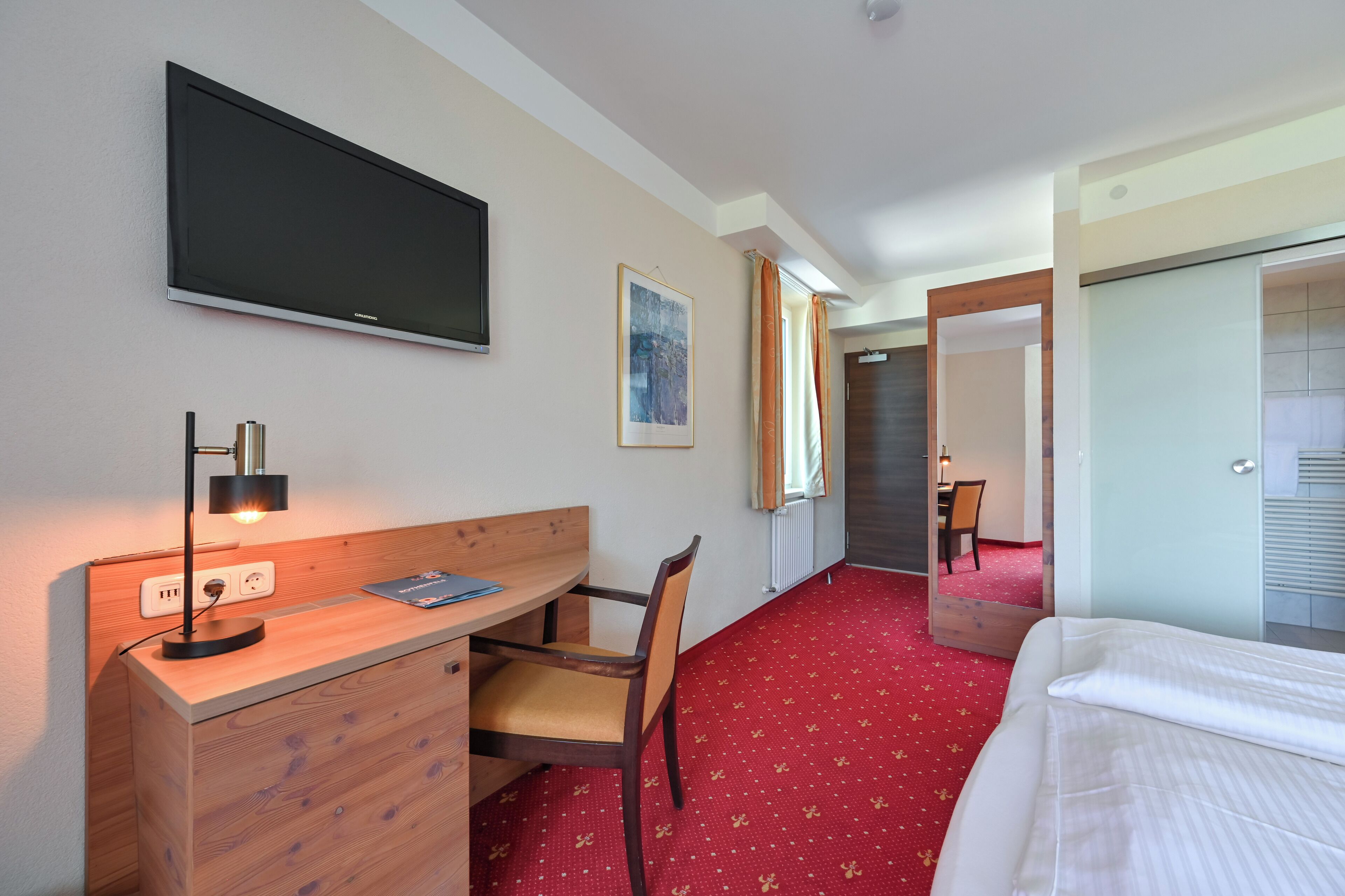 A hotel room with a bed, a desk with a chair, a TV mounted on the wall, and a red carpet.