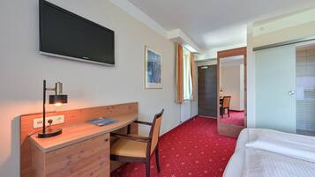 A hotel room with a bed, a desk with a chair, a TV mounted on the wall, and a red carpet.