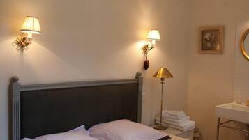 A neatly made bed with a headboard, two wall-mounted lamps, a bedside lamp, a folded towel, a mirror, and a chandelier.