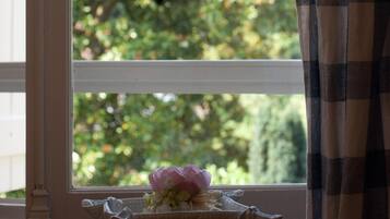 A window with a view of greenery outside, a small basket with flowers on the windowsill, and a checkered curtain partially drawn to the side.
